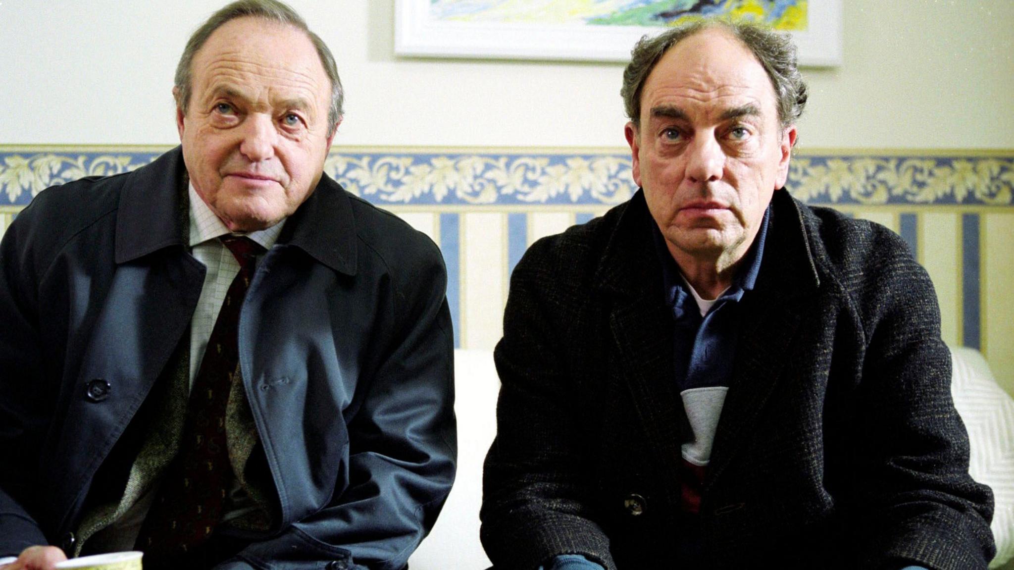 The photo shows actors James Bolam and Alun Armstrong sitting side by side on a sofa. They are holding cups of tea and looking at something off camera. 