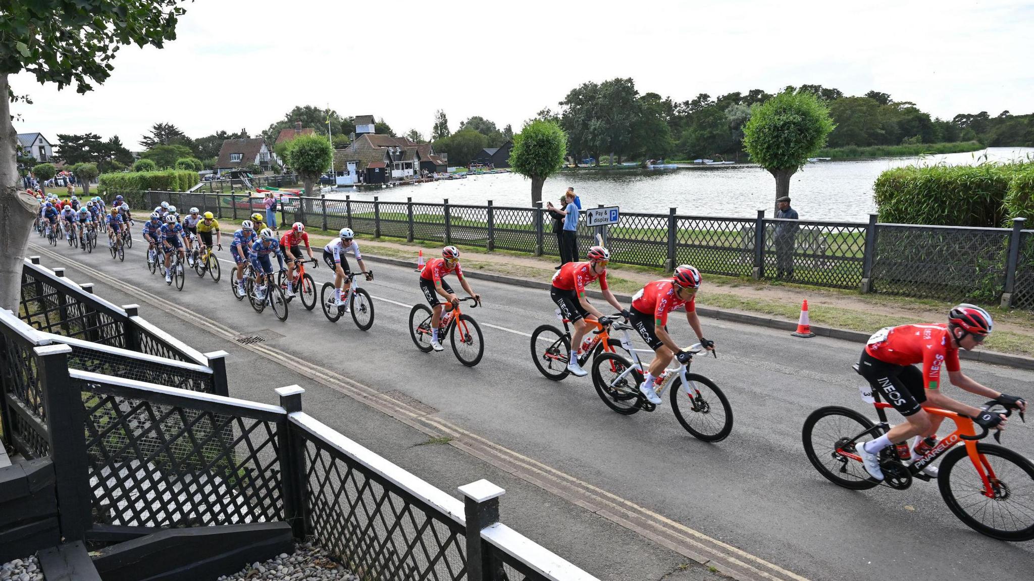 Cyclists race across a bridge over a river. Some spectators watch on from the side of the road.