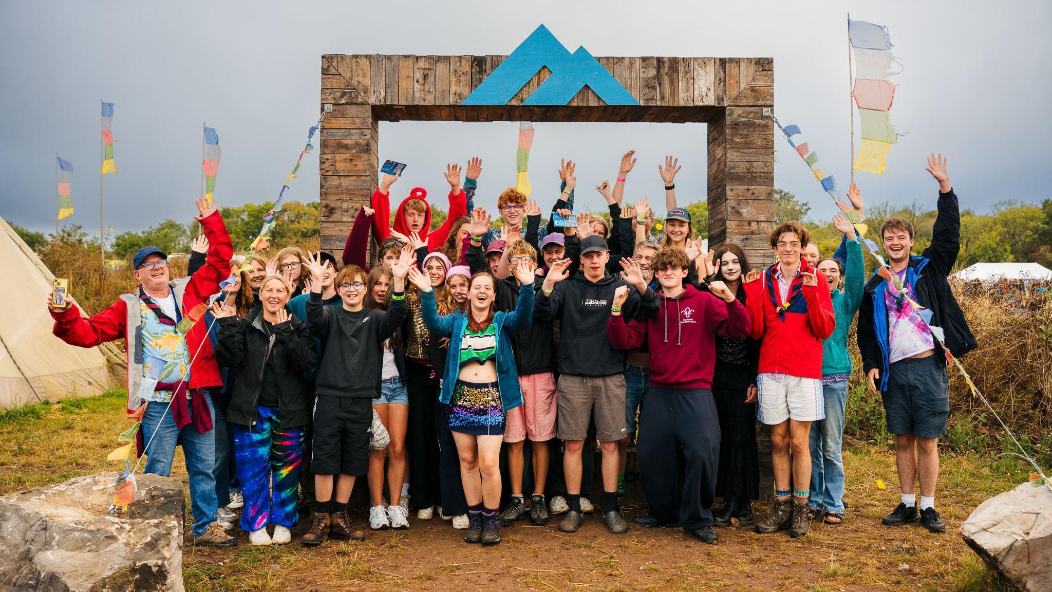 A large group of young people, many of them wearing shorts, smile and wave at the camera at the entrance to the Mendip Adventure Centre. There is a large archway with the centre's logo in blue on top (two triangle shapes). In the background colourful tents and flags are visible
