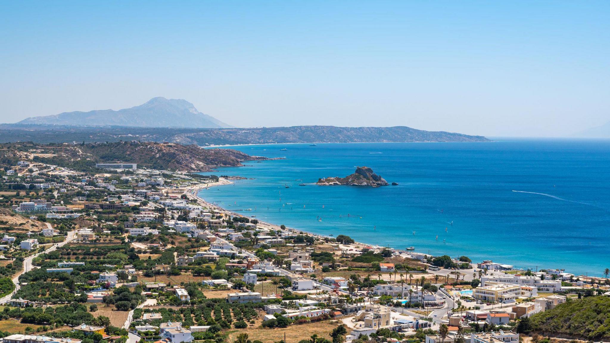 Aerial view of the coastline of Kos, showing blue sea, a thin line of white sandy beach and a village made up of white buildings