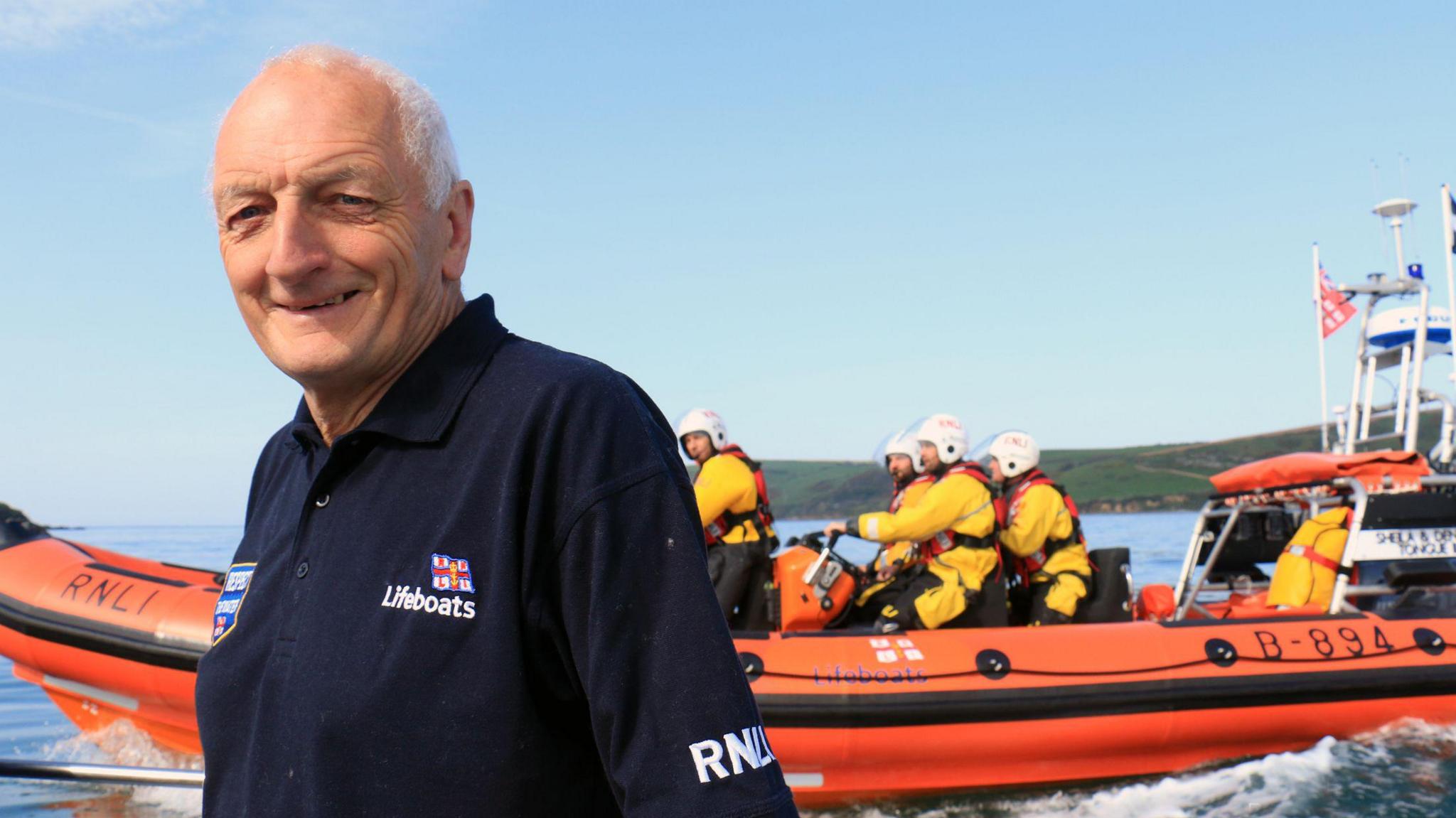 David John Haines standing to the left wearing a navy blue T-shirt with a orange lifeboat and crews on board. He is smiling at the camera and the T-shirt has a RNLI badge on the right. The lifeboat crew are all wearing yellow suits. 