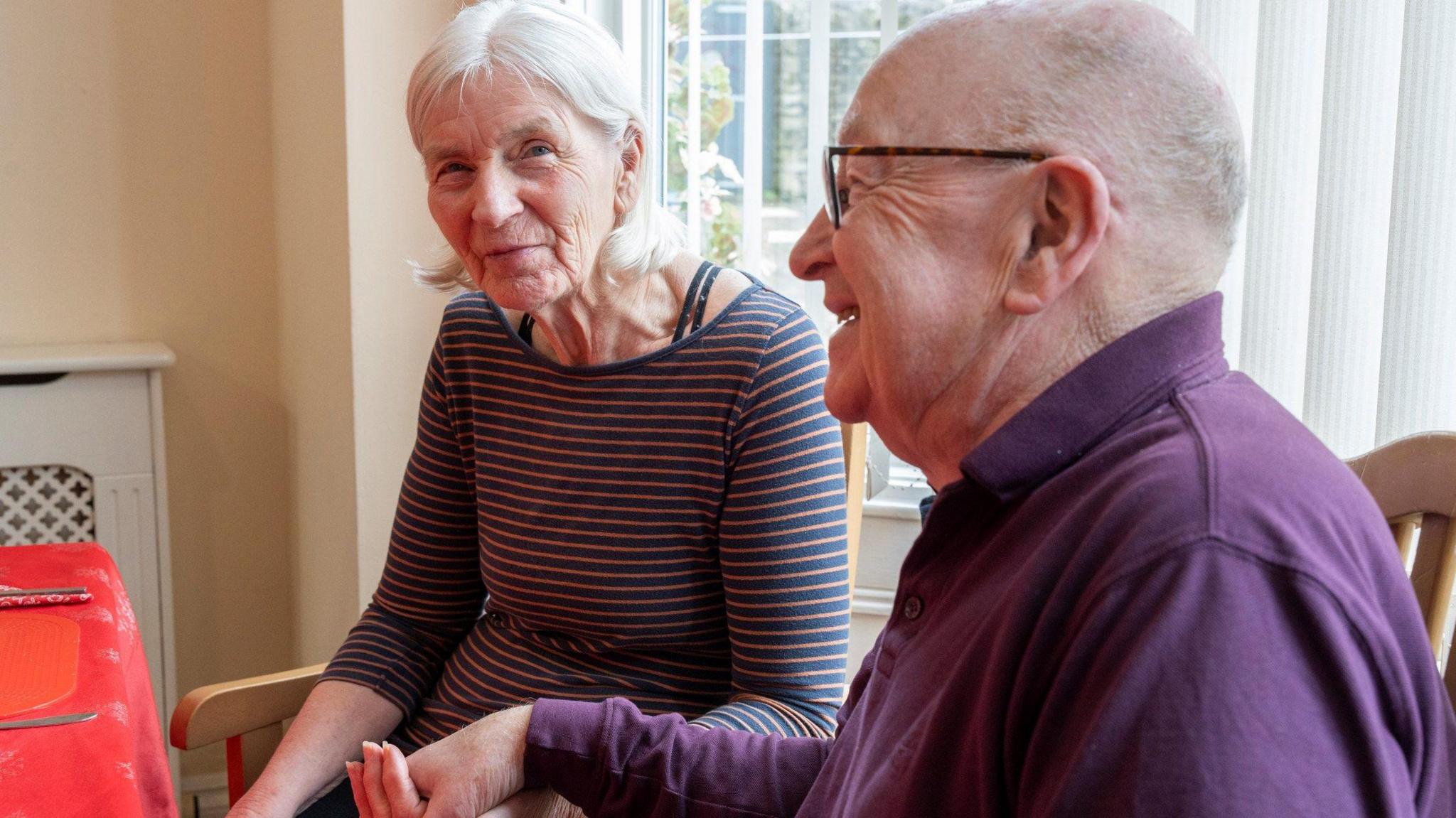 The picture shows Pat and Pete Dalgarno sitting together in a room.  They are holding hands.   Pete is smiling and looking away from the camera.