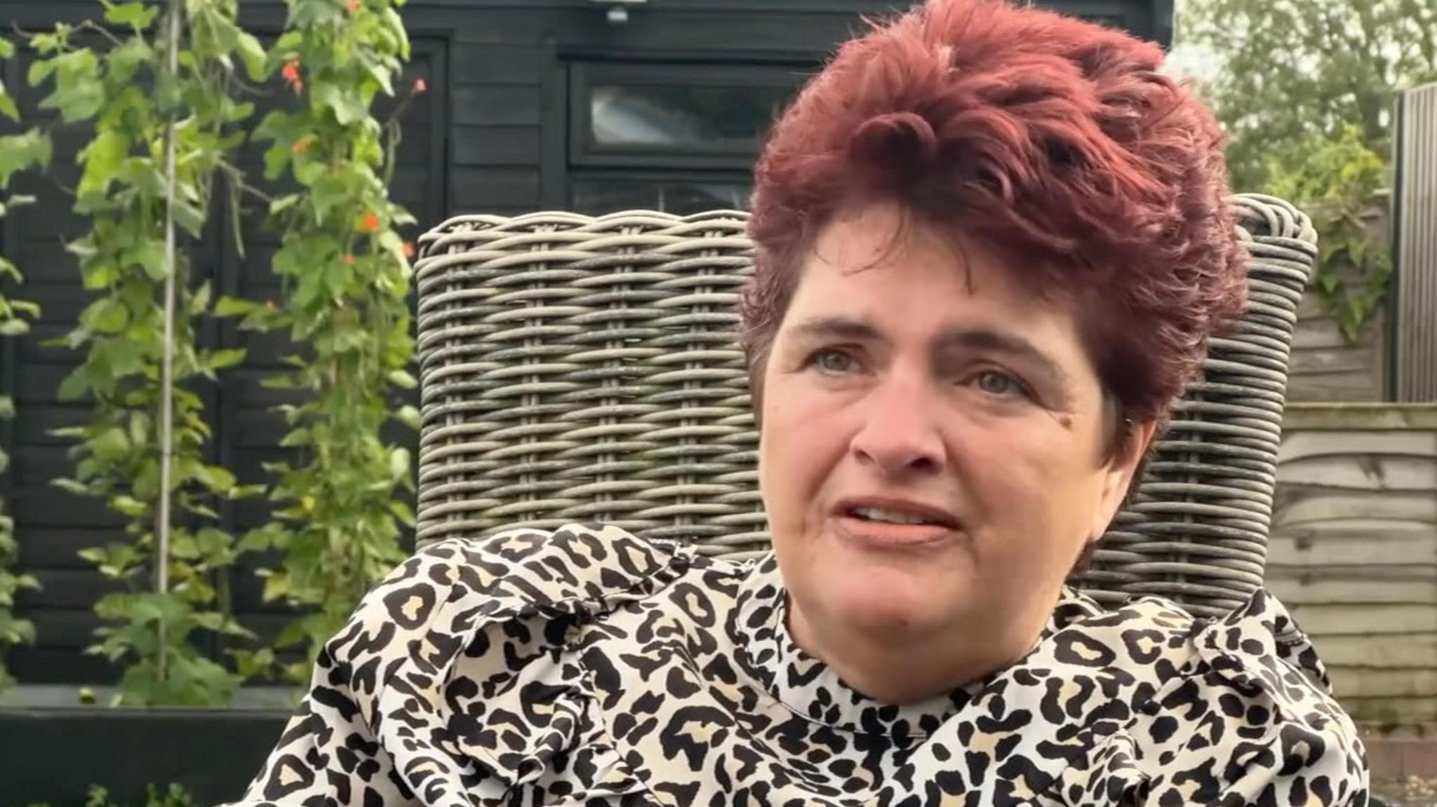 Alison Kingsley has red hair and is wearing a cream and brown-patterned top. She is sitting in a light-green wicker chair in front of a row of tall plants and a black wooden building with a window.