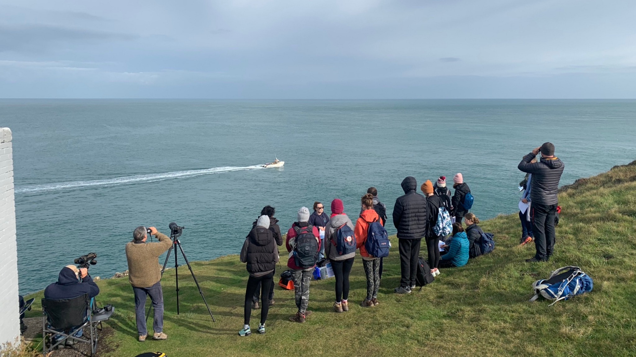 A picture of a group of volunteers completing land-based surveys on the North Pembrokeshire coast. There are about 20 people observing the porpoises from the cliff. Some of them have cameras and binoculars. There is a boat in the sea and it is a sunny day. 