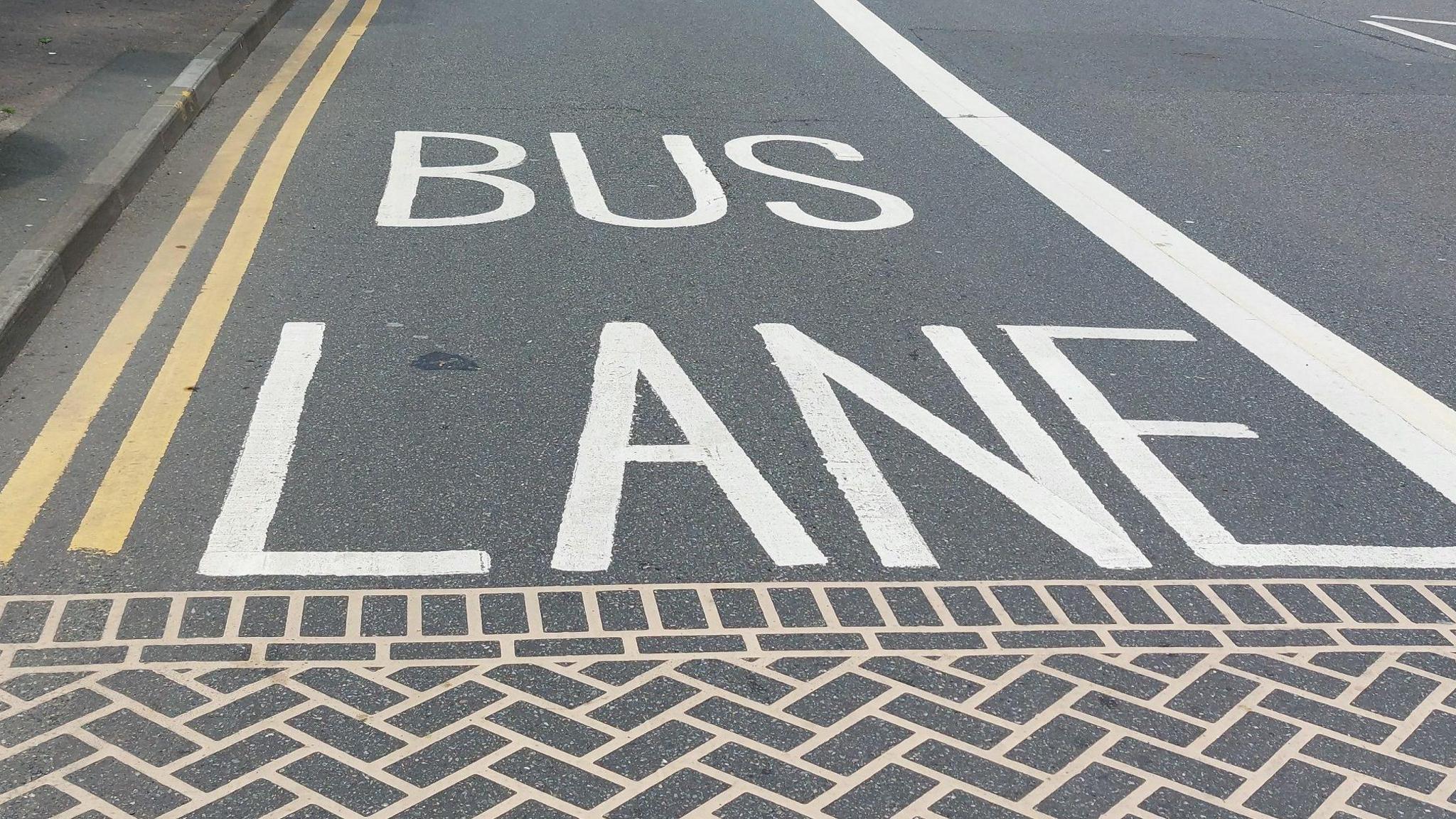 A road, with double yellow lines along the left hand side of it and a white line on the right hand side, with "BUS LANE" written in the middle of it, with what appears to be patterns like a brick path along the bottom