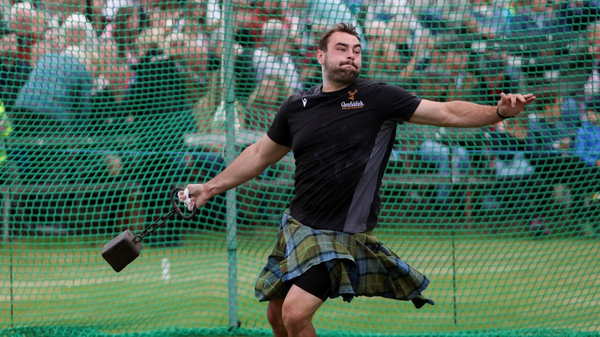 A man is taking part in the hammer throw with a grimace on his face. He is wearing a black T-shirt and a tartan kilt. He is about to throw a hammer connected to a handle via a chain. There is green safety netting behind him and spectators further back.