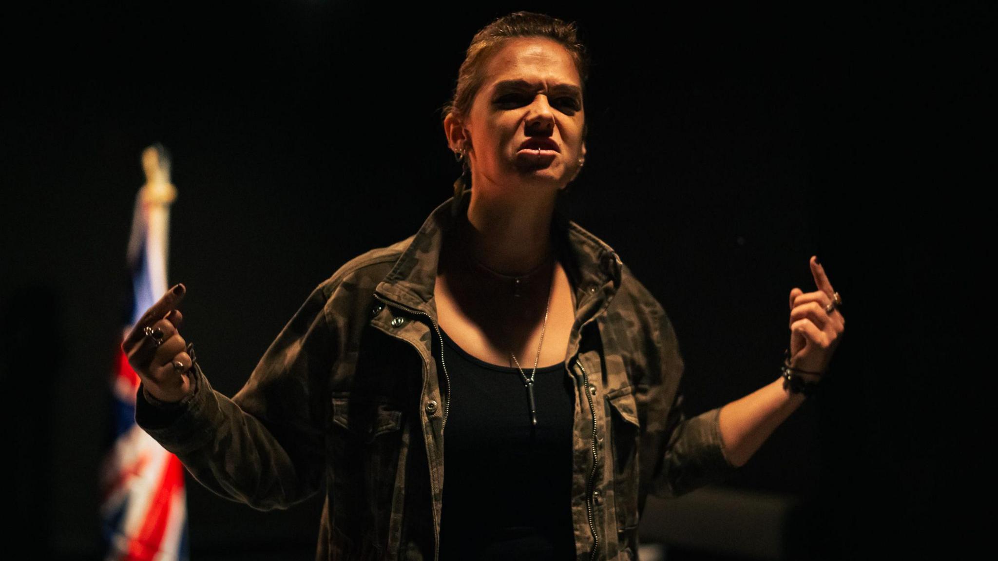 An actor in a black top and brown jacket stands on a dark stage with a Union flag in the background, gesturing and pulling an angry face, in the new play At Last.