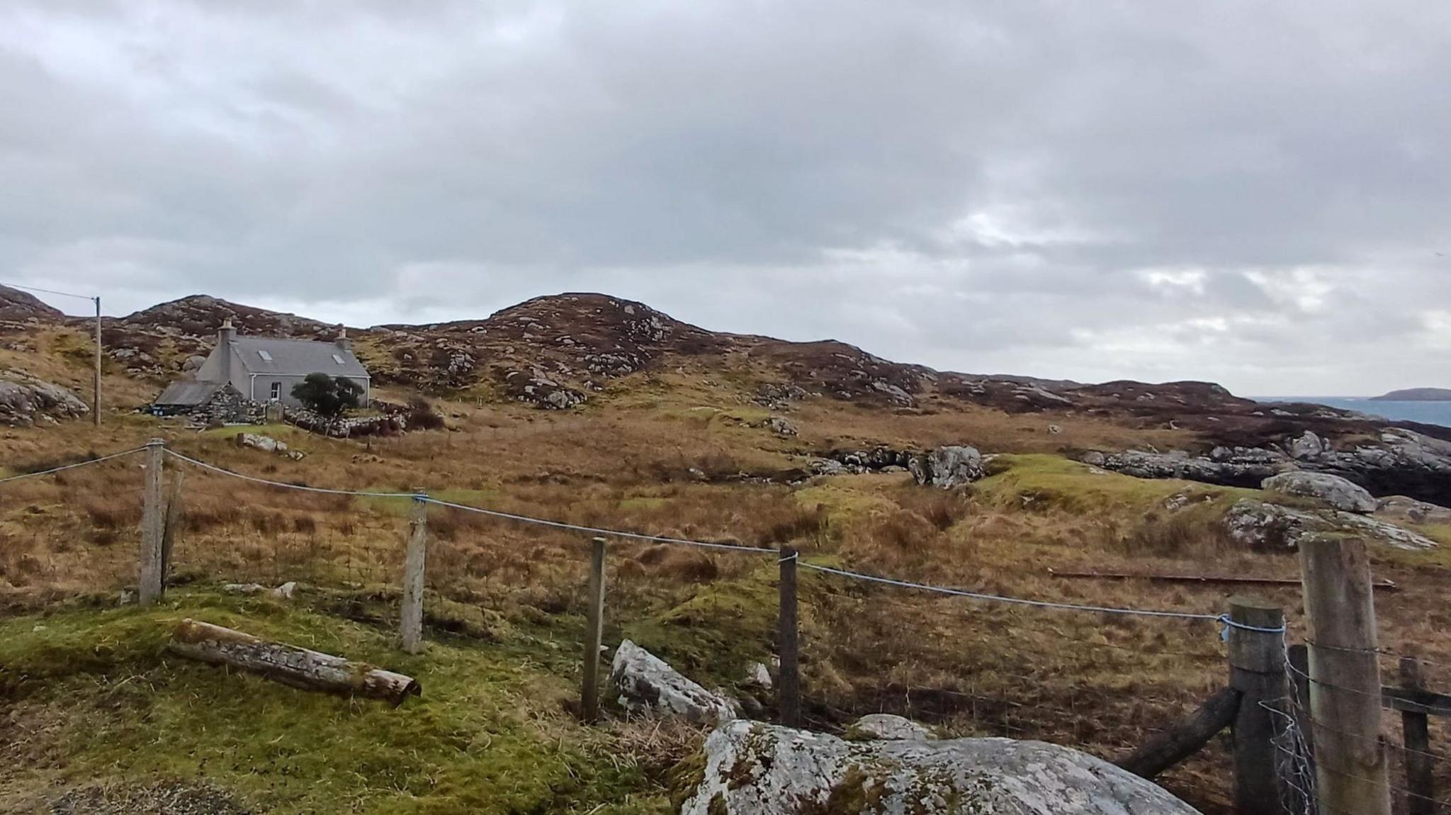 The small, single-storey house sits in a landscape of moor and bare rocks. There is a wire fence in the foreground.