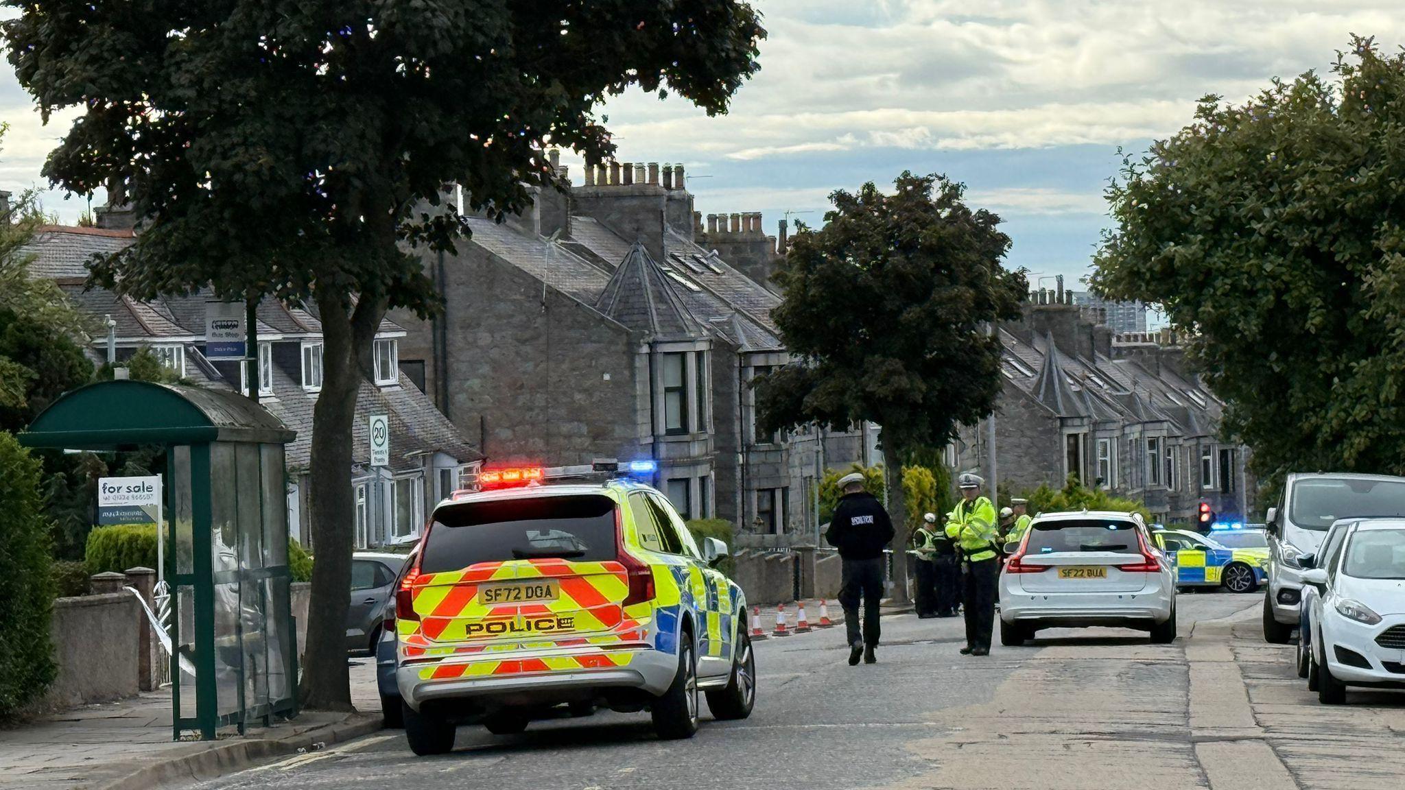 Police officers and police cars in a residential street with stone houses and trees. The patrol car int he foreground is parked next to a bus stop shelter and has its lights on.  