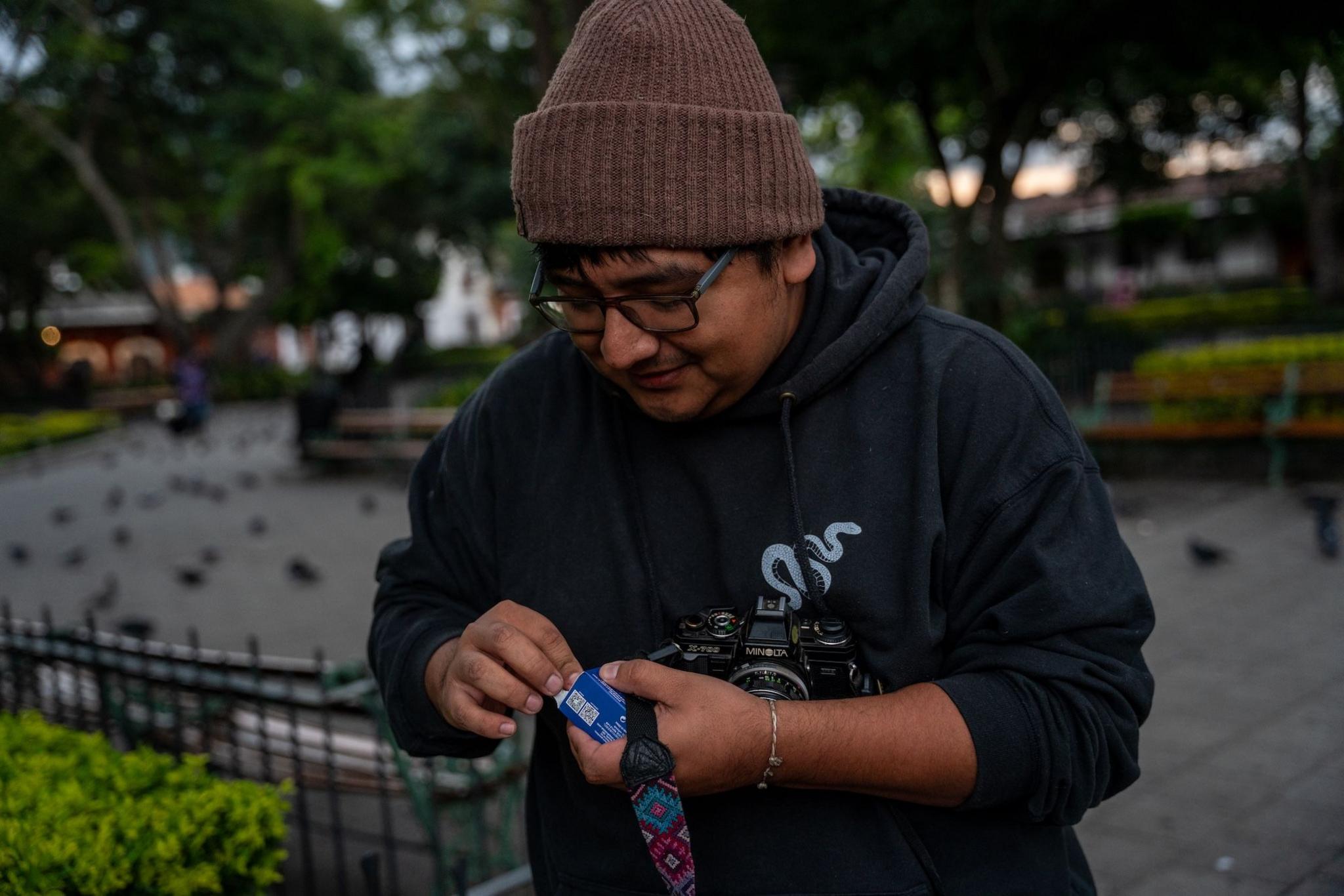 Ronald Ottoniel, wearing a woolly hat and thick-rimmed glasses, is loading his new film roll into his camera ay a square in Antigua. Pidgeons can be seen on the ground in the background.