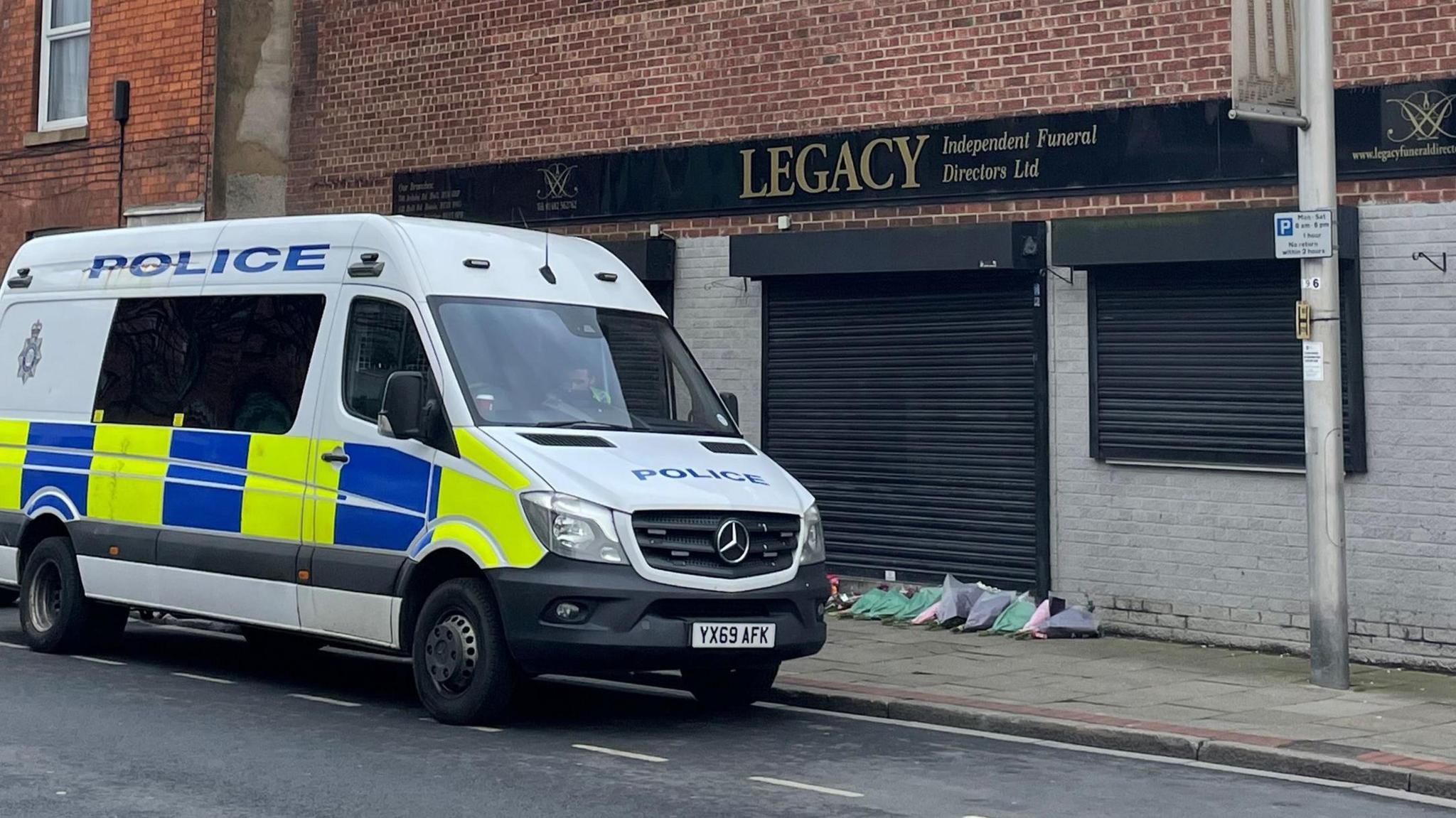 A white police van, with yellow and blue decor, outside the premises of Legacy Independent Funeral Directors, in Hessle Road, Hull. The large, red-brick building has black shutters, a flat roof and a black and gold "Legacy" sign. White graffiti covers part of the front wall, with more graffiti on a neighbouring roof.