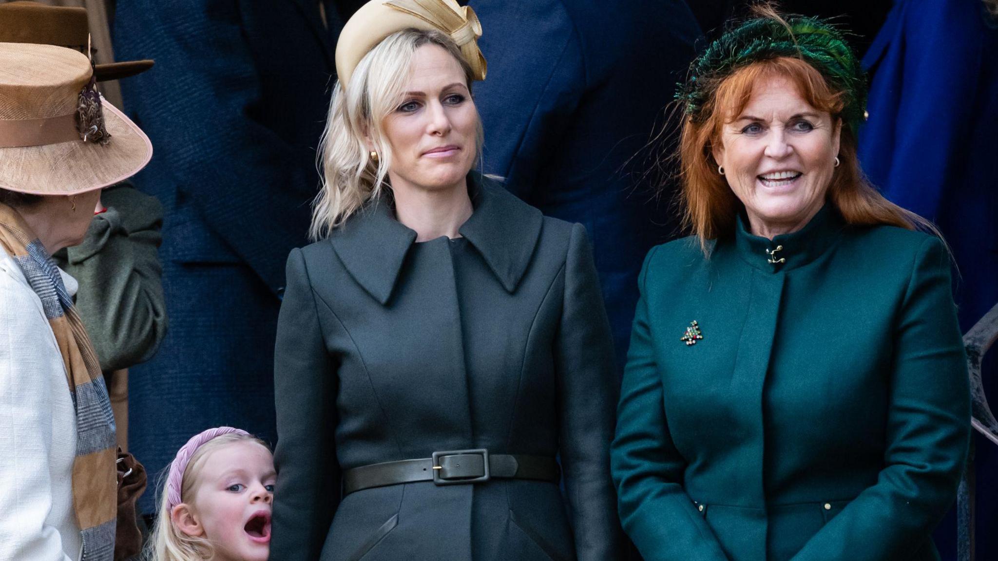 Two women stand together - one is wearing a bottle green, the other a navy  coat. One wears an elaborate head band displaying bottle green feathers while the other woman sports a beige hat with a bow. Another female can be seen wearing a taupe coloured hat. A young child stands close to her mother and has a pink head band. 