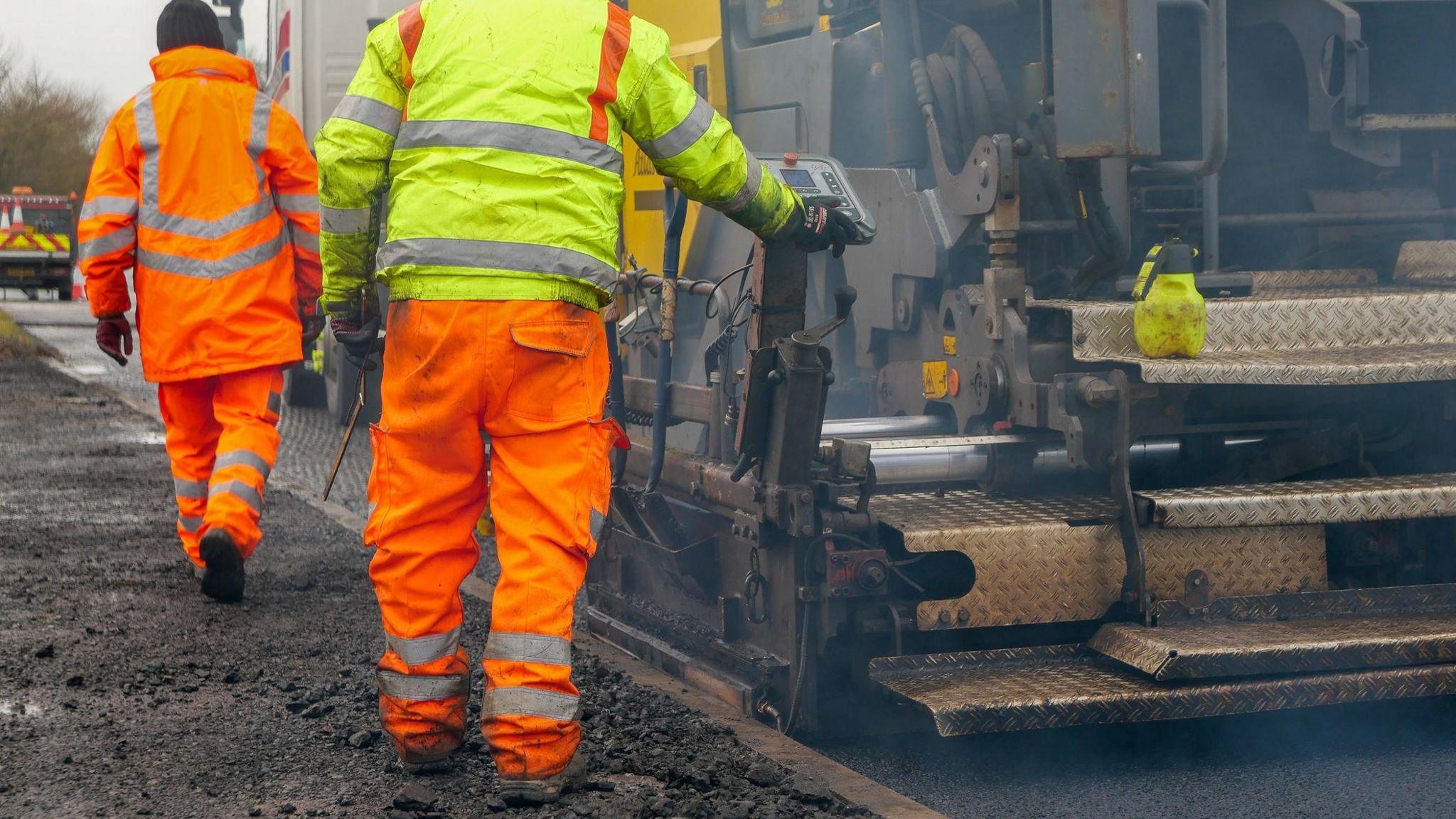 Two men in high-vis jackets and trousers on a construction site. They are facing away from the camera and stood on a gravelly path beside a large van.