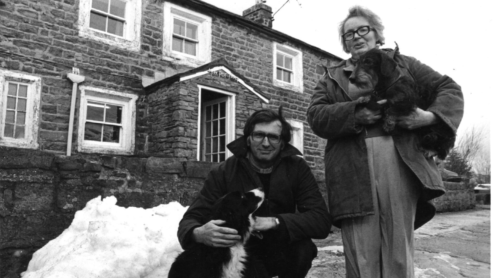 A husband and wife are standing in front of their brick house in Snaizeholme, near Hawes, in a black-and-white picture. The man is squatting down embracing a Collie, and the woman is standing up holdind another dog in her arms. 