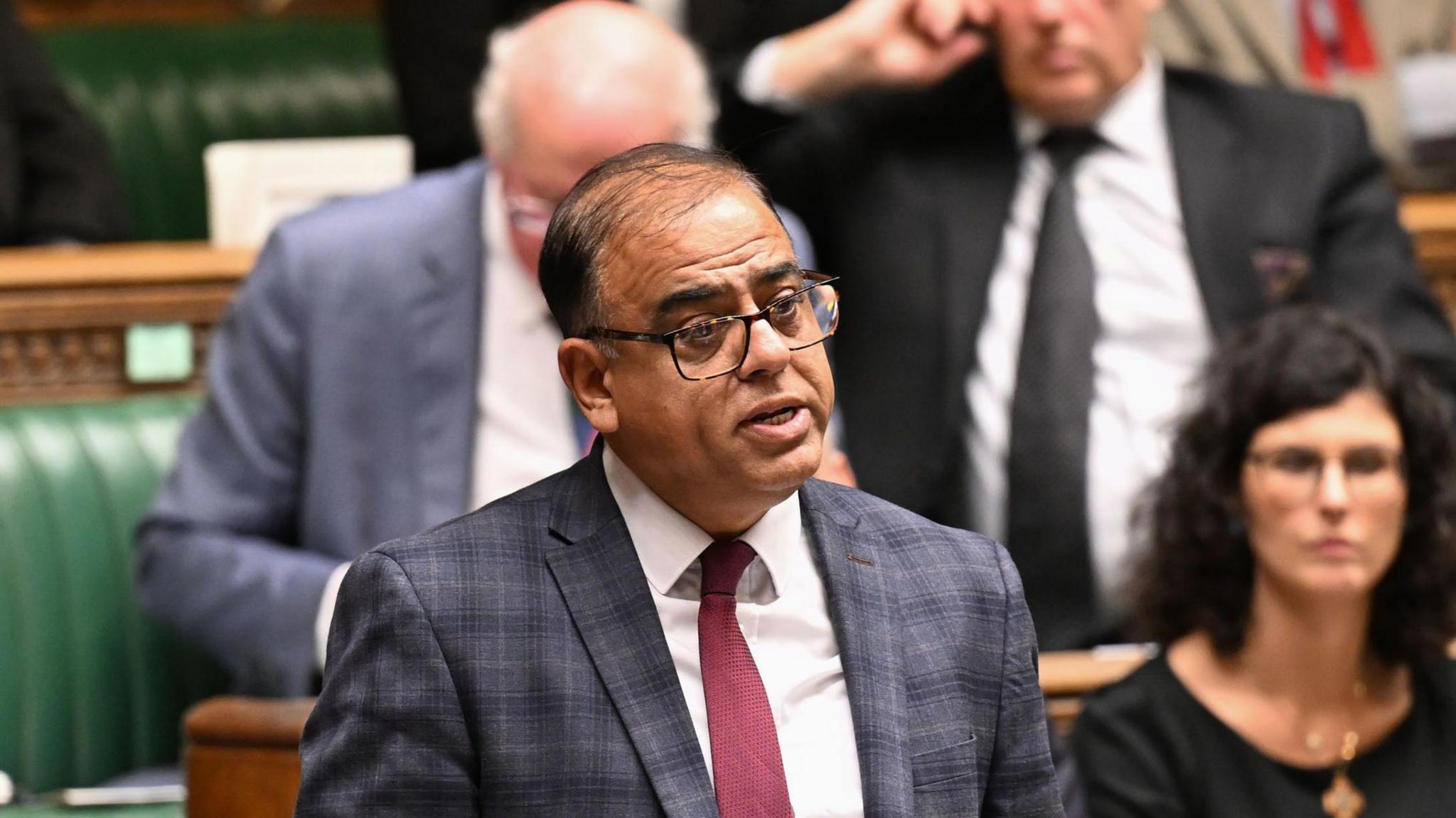 Mohammad Yasin is pictured speaking in the House of Commons. He has dark thin hair and wears a chequered blue and grey suit with a white shirt underneath and a red tie. Other MPs are pictured sitting and listening behind him.