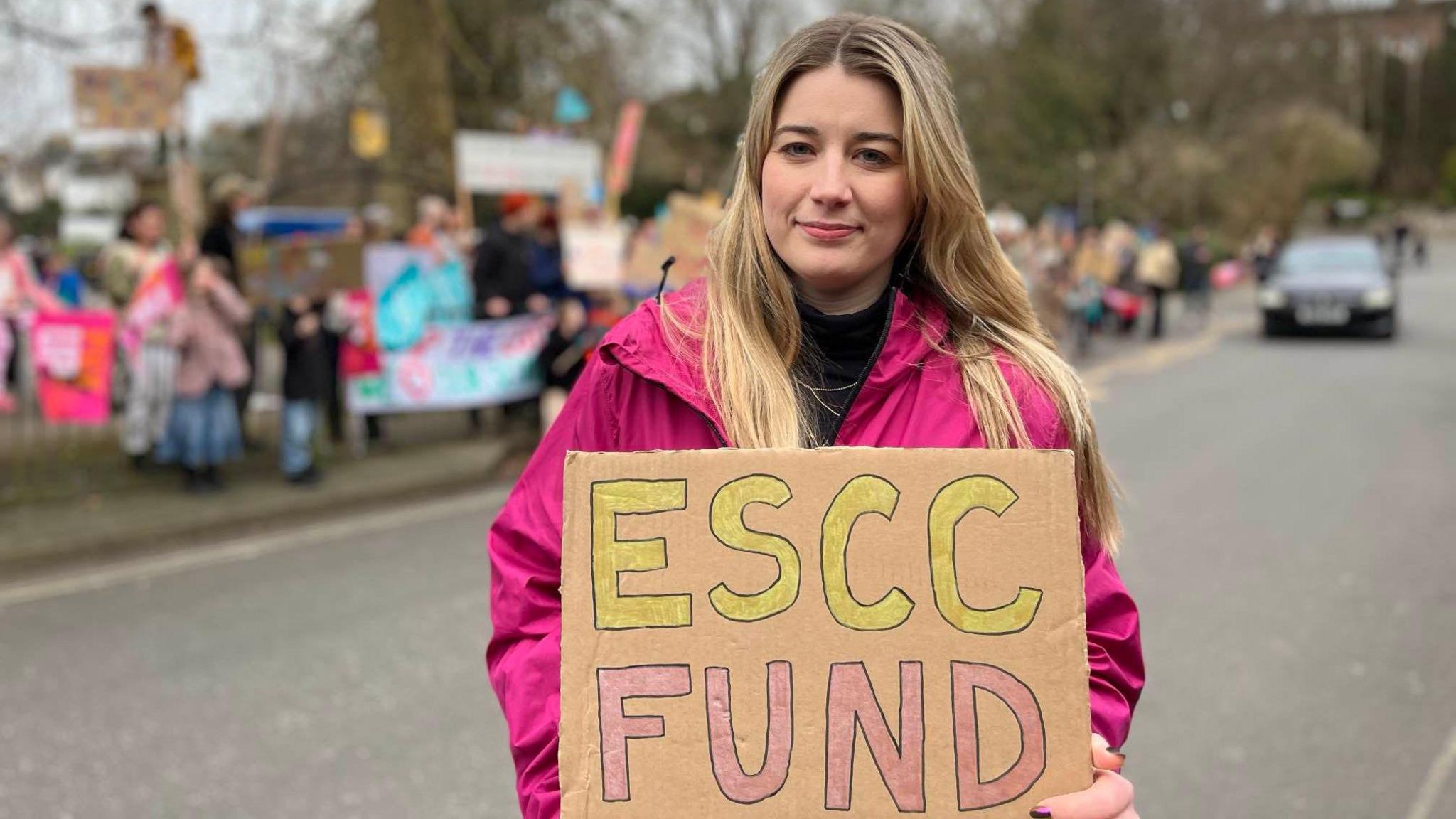 A woman in a pink coat holding a sign reading "ESCC Fund"