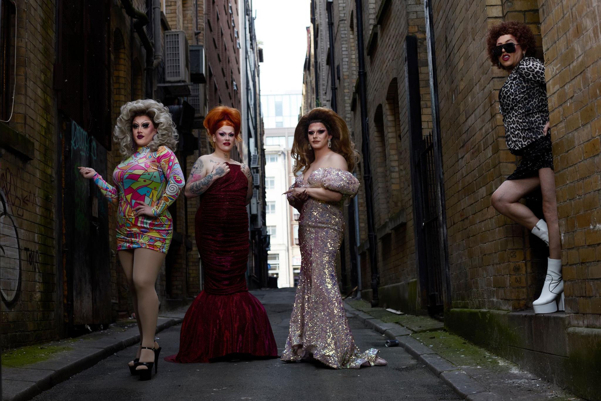 Four drag queens pose in an alley way, one wears a rainbow mini dress, another wears a red velvet floor-length gown, the third wears a sparkly gown while the fourth stands in a door way wearing a mini skirt, a leopard print blouse and white go go boots