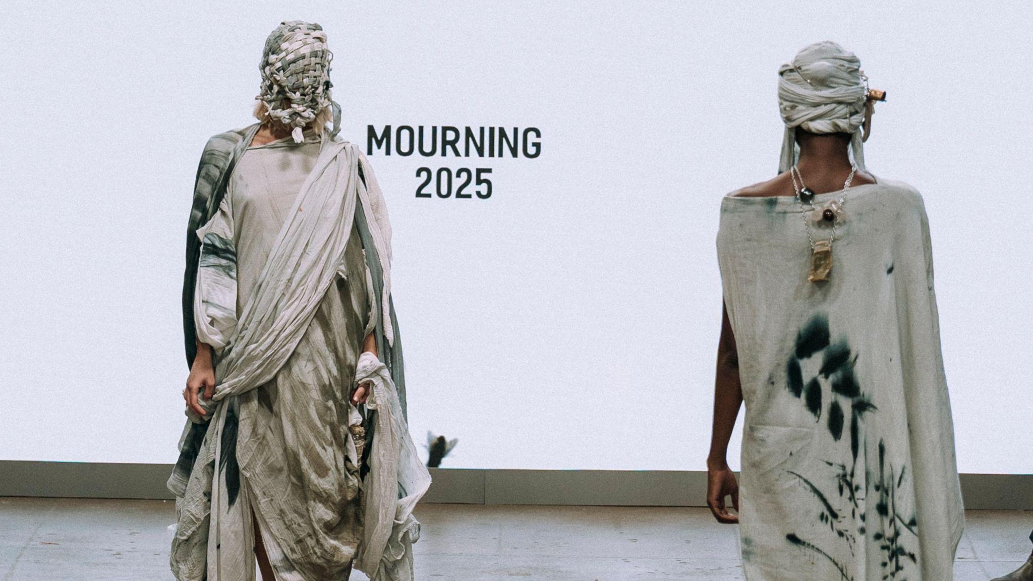 Two models walking along a catwalk in opposite directions. They are both wearing long cloaks made from fabrics stained by the weather. Both have rags wrapped around their head and faces. 