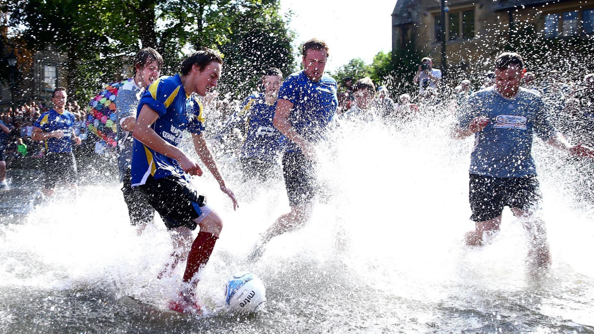 Footballers playing a match while water is kicked up around them. There are peopel wathcing and taking photos in the background.