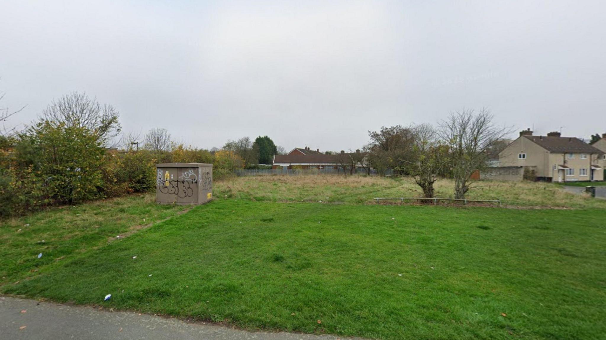A grassy strip of land with two small trees and bushes on it. Some houses are visible in the far right of the picture.