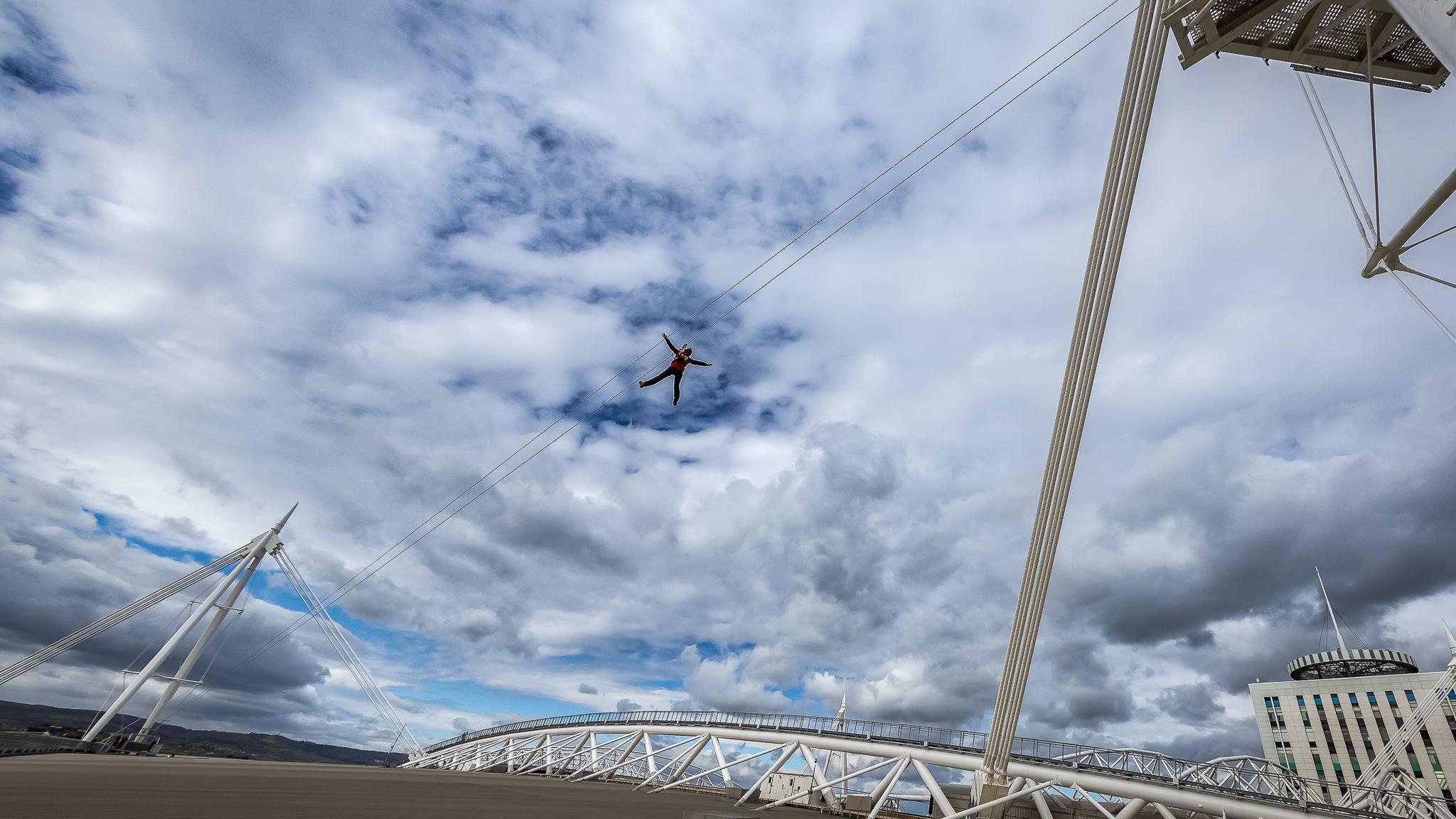 The roof of the Principality Stadium in Cardiff looking out over the city. Its white mast-shaped pillars are strung with a wire that hold a person zipping through the air with their arms and lets spread out. There is a blue sky a clouds in the background and a white building with a spike on top.