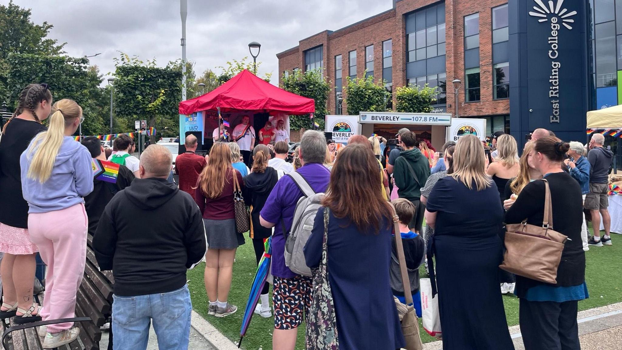 Crowds of people with their backs to the camera watching a performance on a small stage with a red canopy. They are stood on a section of grass which is surrounded by a path. The sky is overcast. 