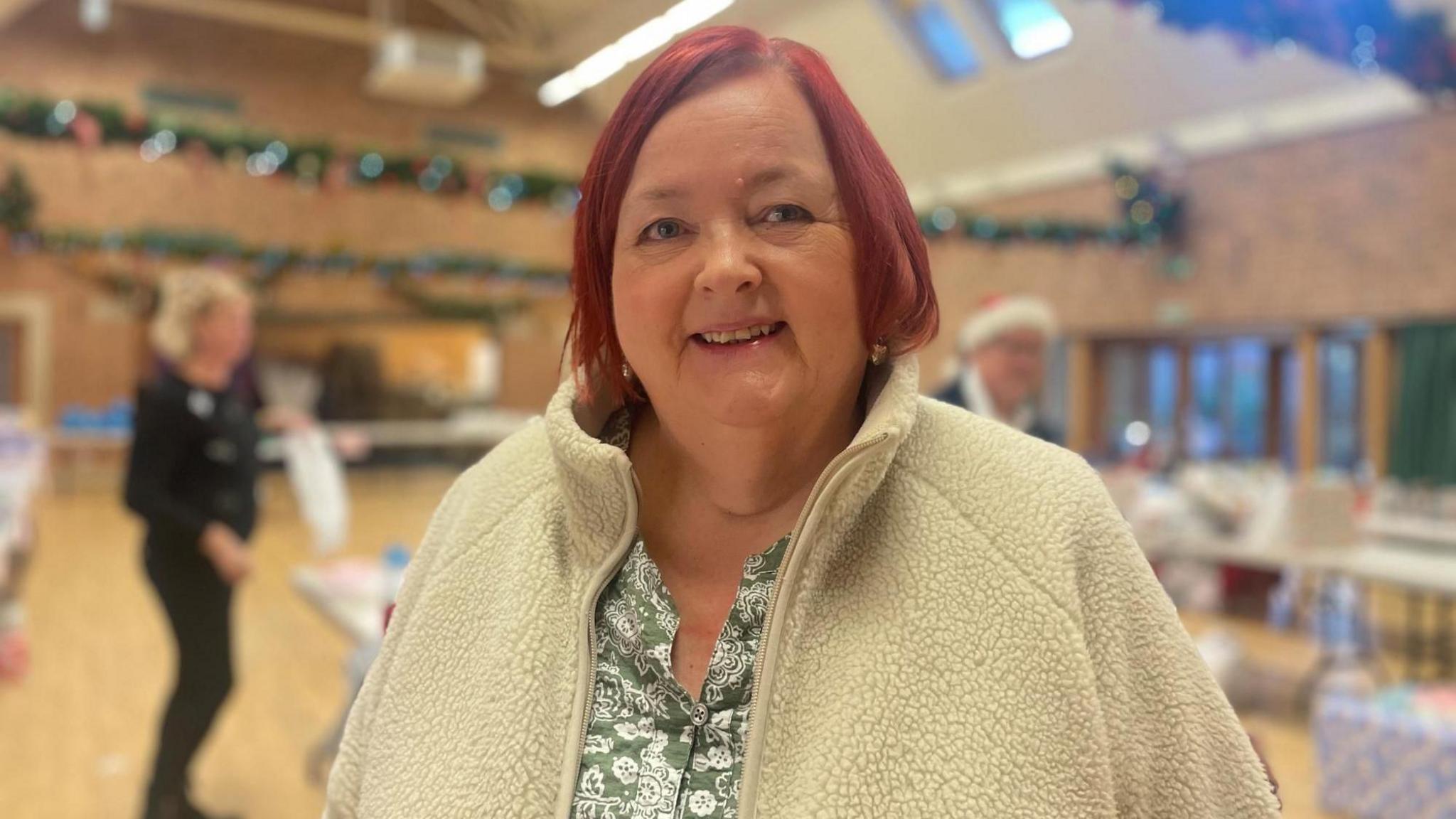 A woman with bobbed red hair and a white fluffy fleece smiles at the camera