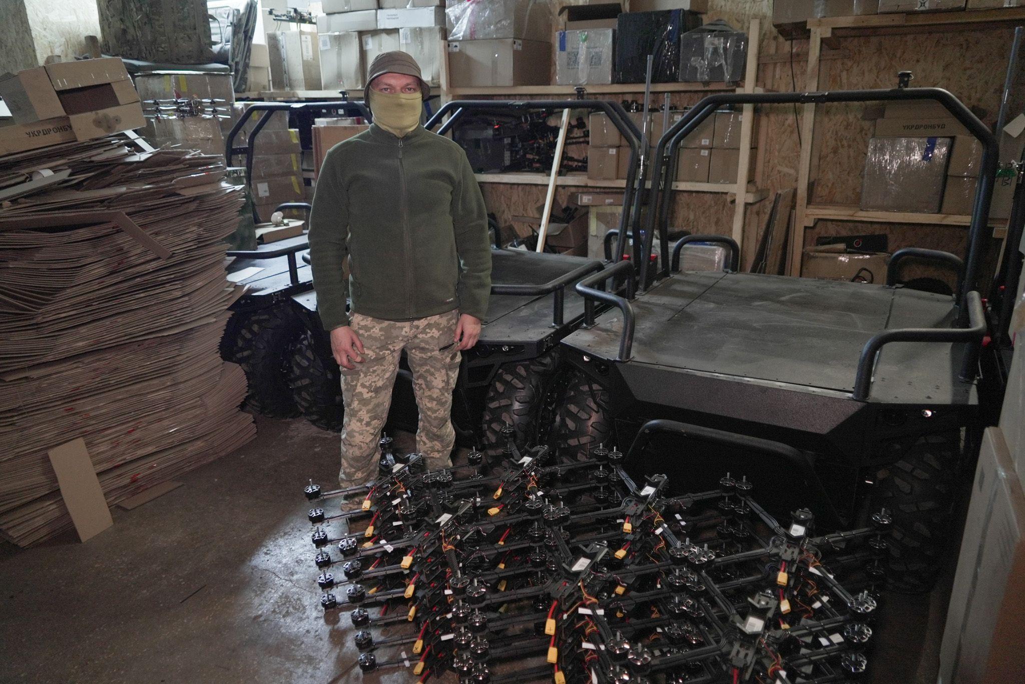 A man in a warehouse standing with a stack of aerial drones and three land drones. He is wearing military fatigues and and has his face covered with a green snood.
