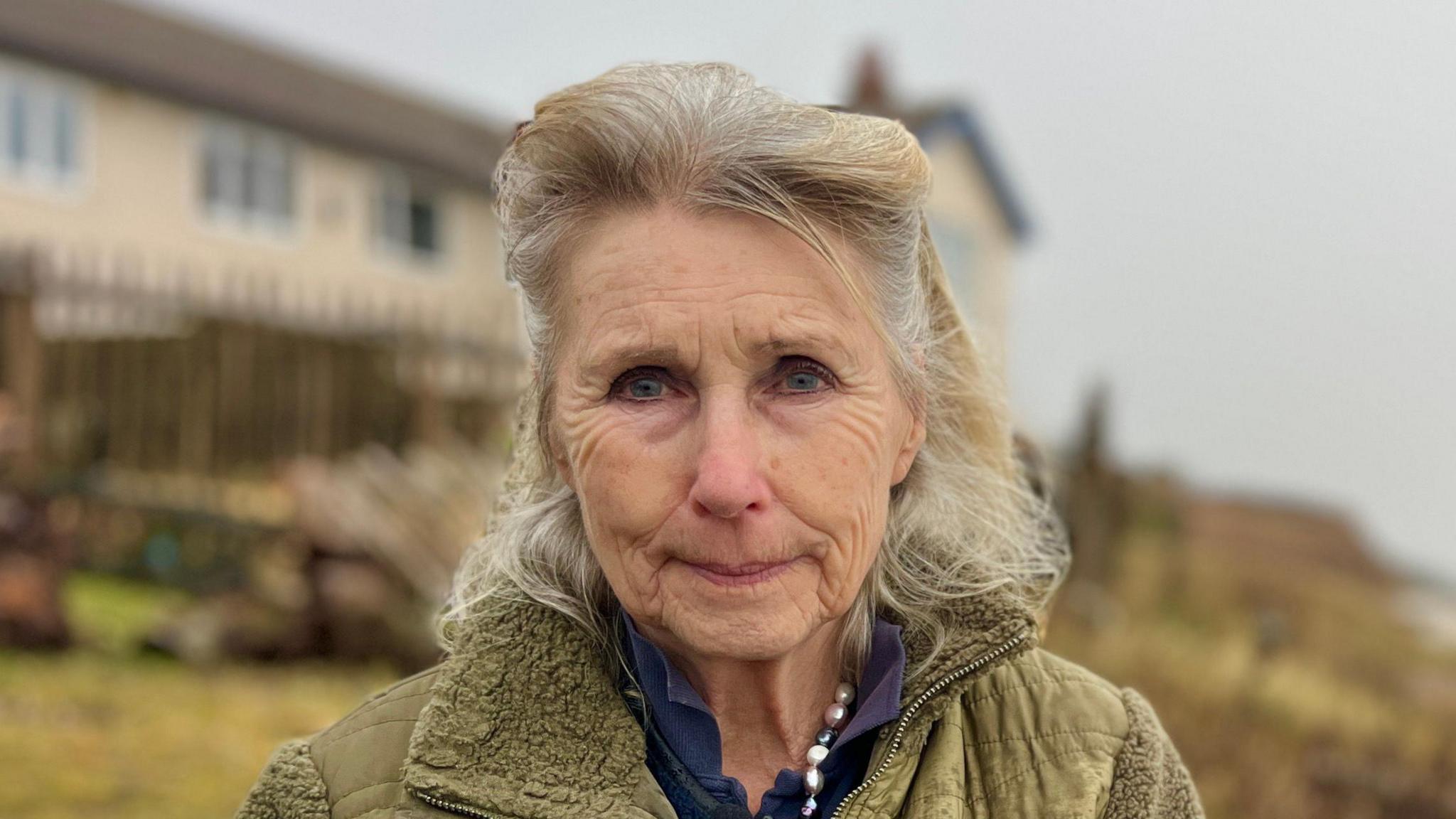 Hilary Lightfoot smiles at the camera. She has short grey and blonde hair and is wearing a green coat with a blue top underneath. Behind her, coastal homes can be seen.