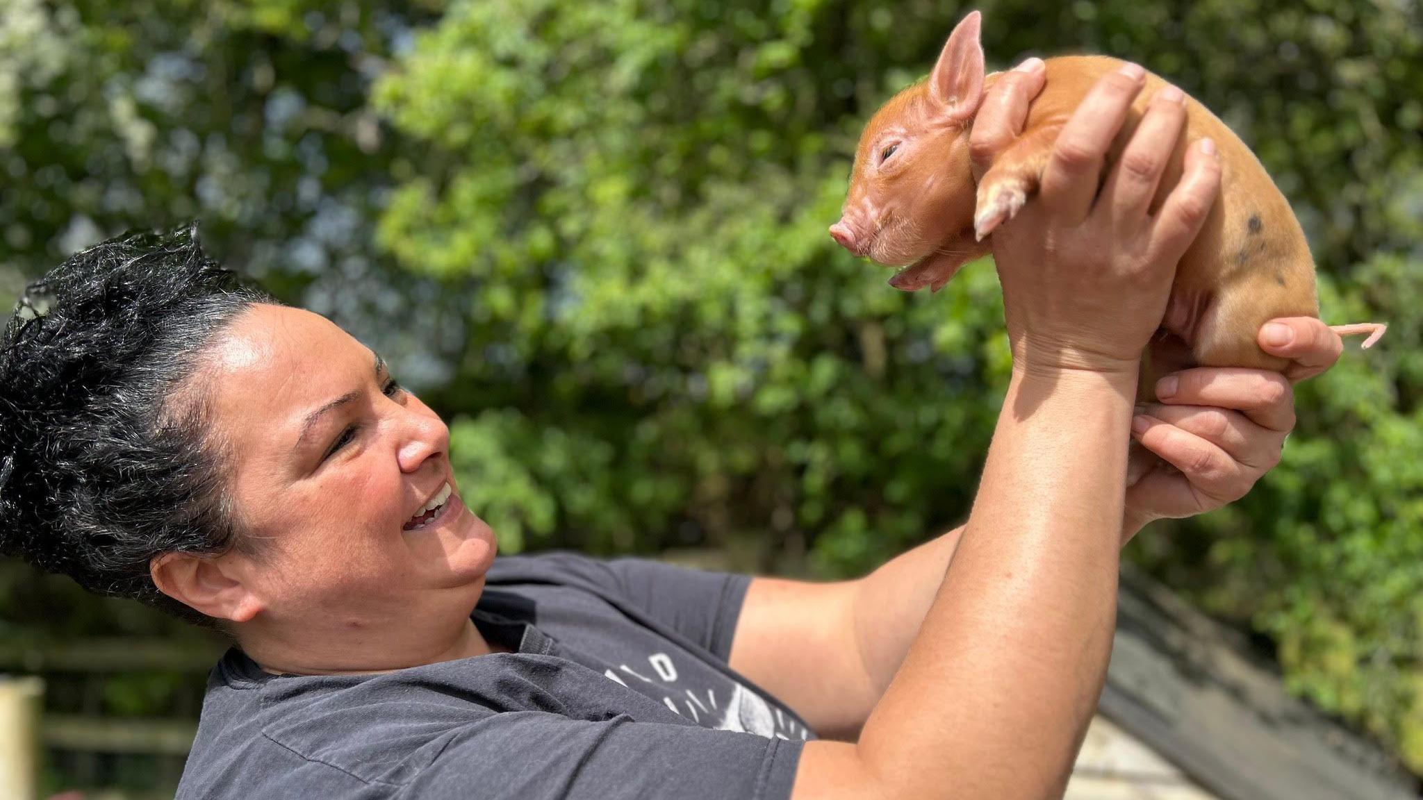 Olivia Mikhail has dark hair and is holding a micro pig up in the air. She is smiling.