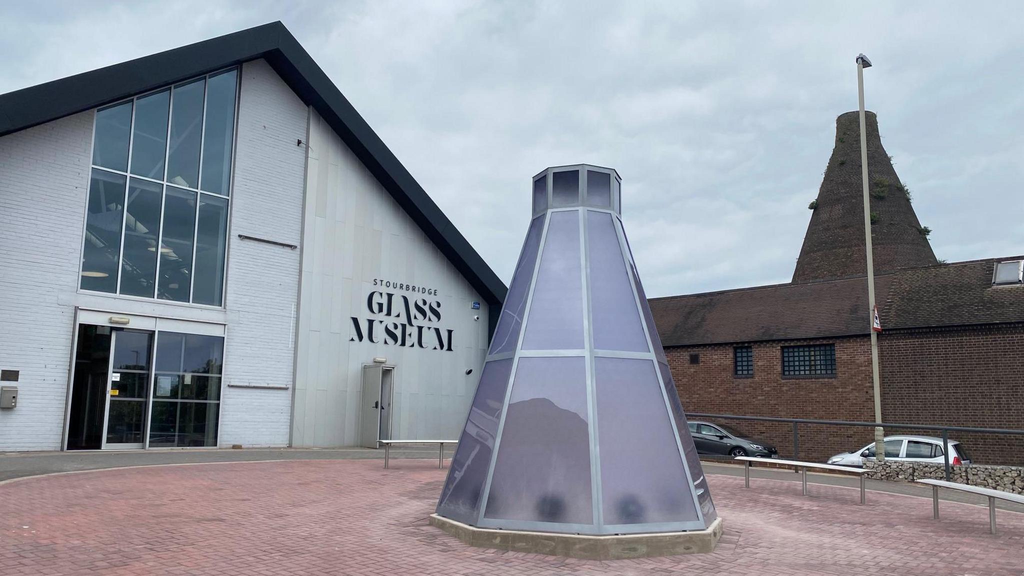 The Stourbridge Glass Museum. It is a modern-looking building. There is a sculpture outside. A sign can be seen on the front.