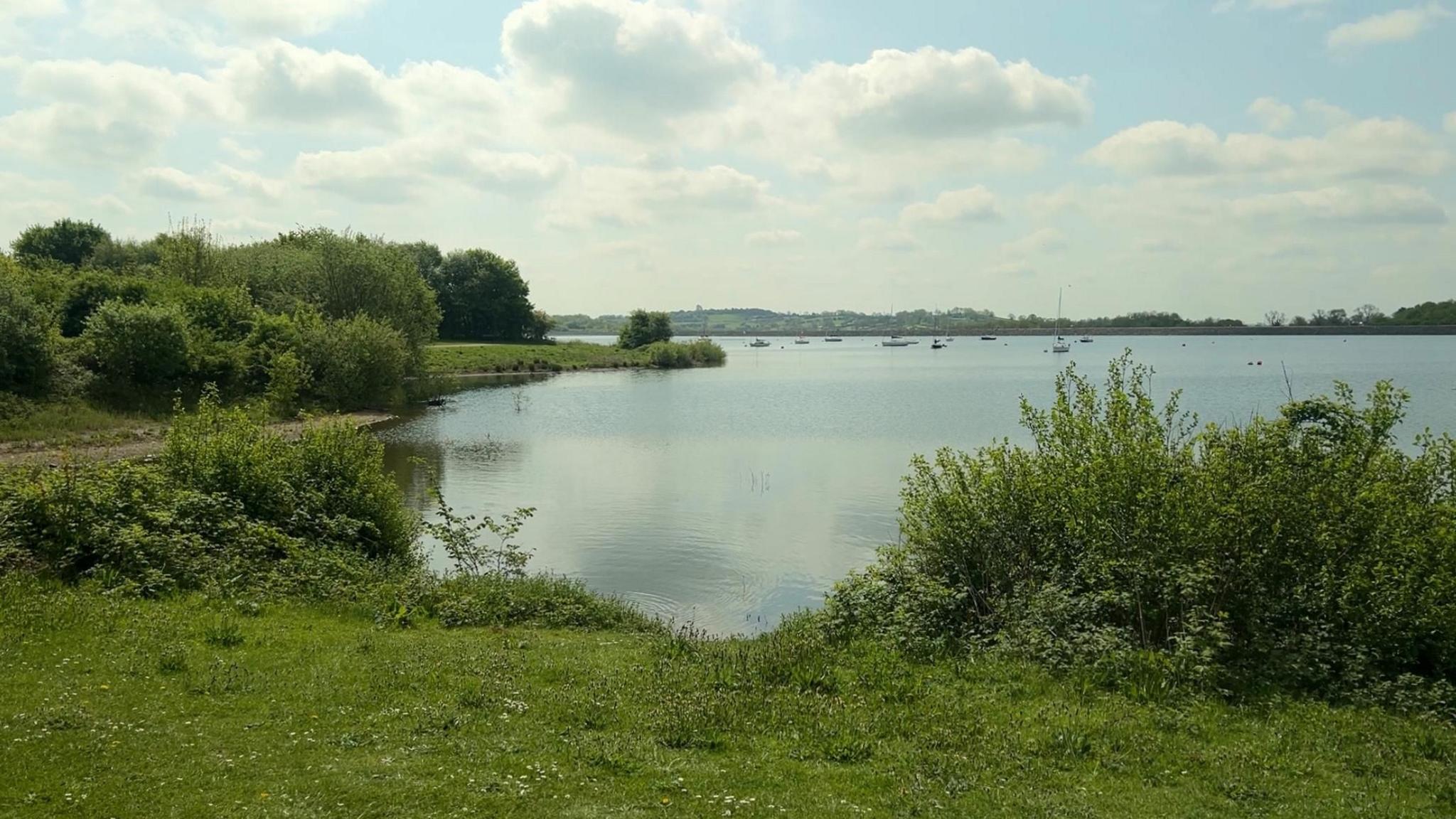 Carsington Water reservoir, which is owned by Severn Trent. The reservoir is a wide expanse of water with some boats visible in the far distance. The banks of the reservoir, with green grass and bushes, can be seen closer to the camera.