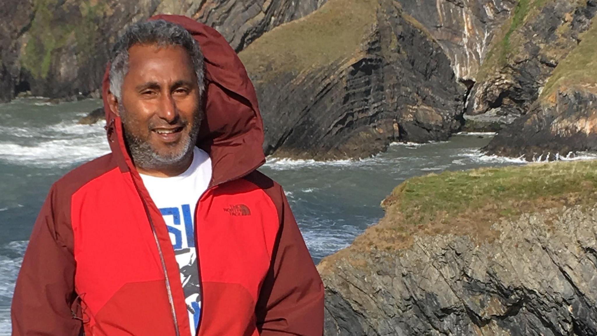 Dr Kuppan is wearing a red coat and white t-shirt, while standing on a wild-looking coastline. The wind is blowing his hood up. There are granite cliffs and rough seas behind him.