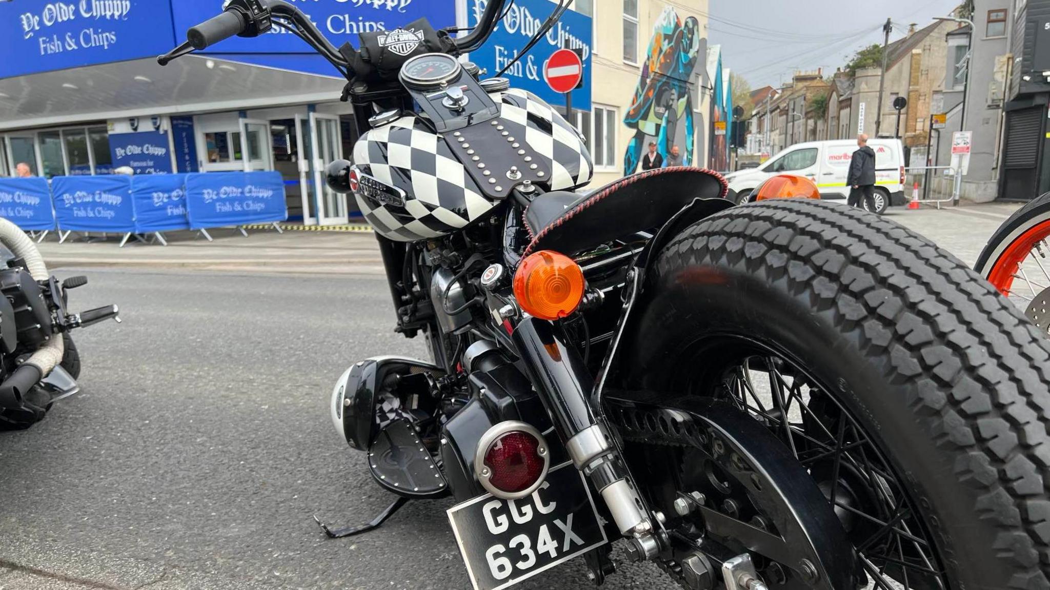 A closer look of Mrs Gelsthorpe's Harley Davidson from behind. The bike's petrol tank has been decorated in white and black diamonds. It has a black leather seat and black handlebars. 