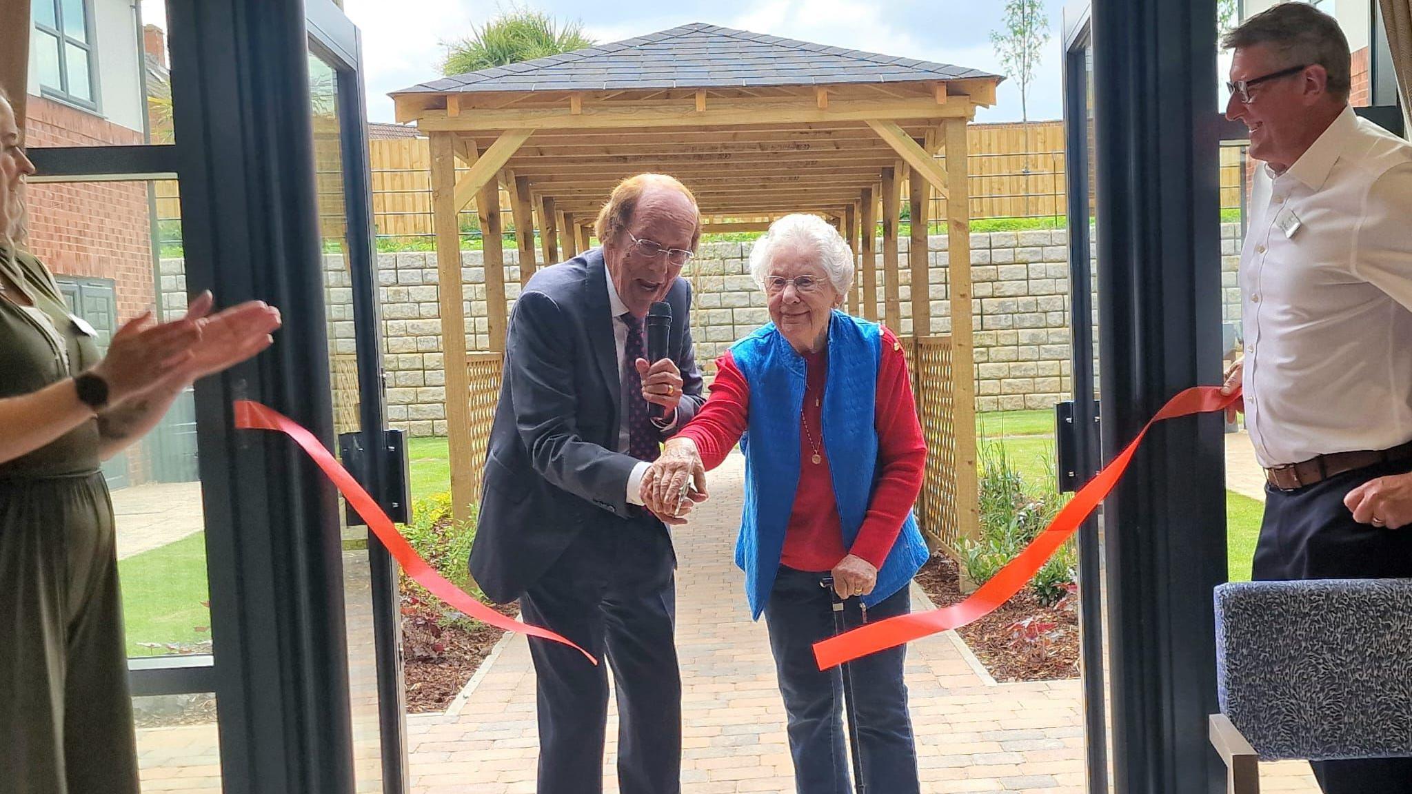 Fred Dinenage MBE together with an older woman have cut a red ribbon in half and are now walking through the entrance of a building towards the camera. A woman to the left is clapping and a man in a suit on the right is smiling.