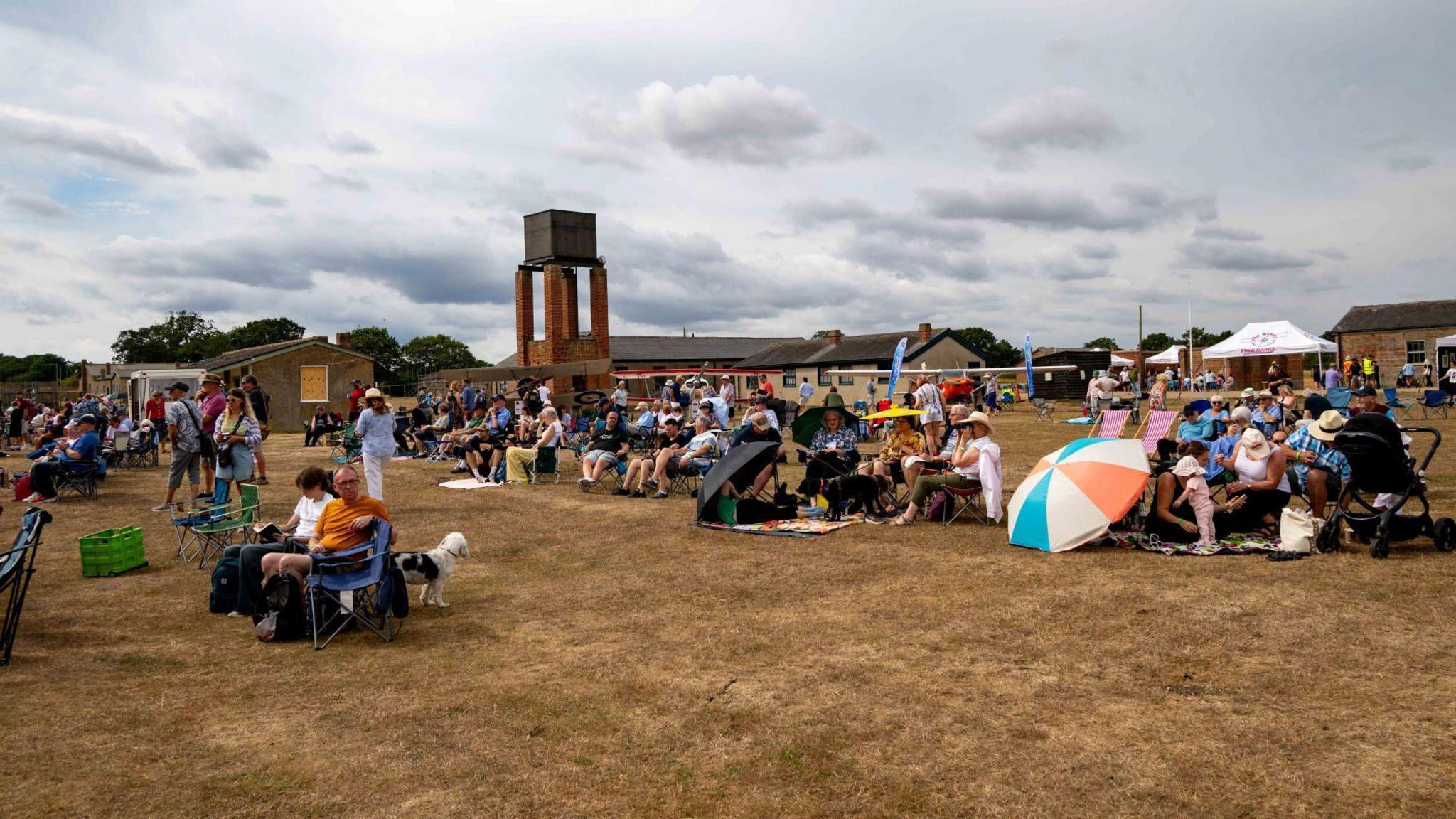 People sitting on grass that looks quite yellow. They have deck chairs and umbrellas. There are about 100 people in the shot.