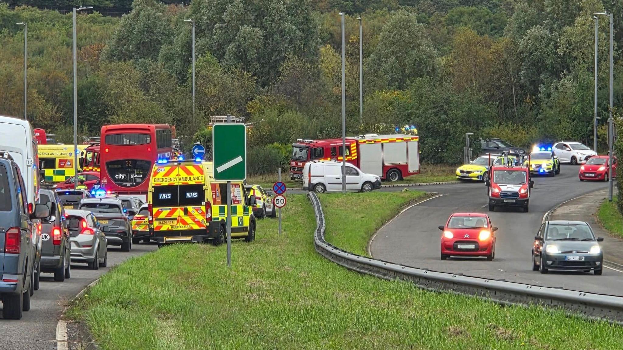 Traffic leading up to Devoran roundabout is at standstill. An ambulance, a fire engine and two police cars all with blue lights flashing are making their way around the roundabout while an ambulance and police car are driving on the verge to get to the scene. The opposite lane has a couple of cars travelling freely. There are trees in the background and a grassy verge in the foreground.