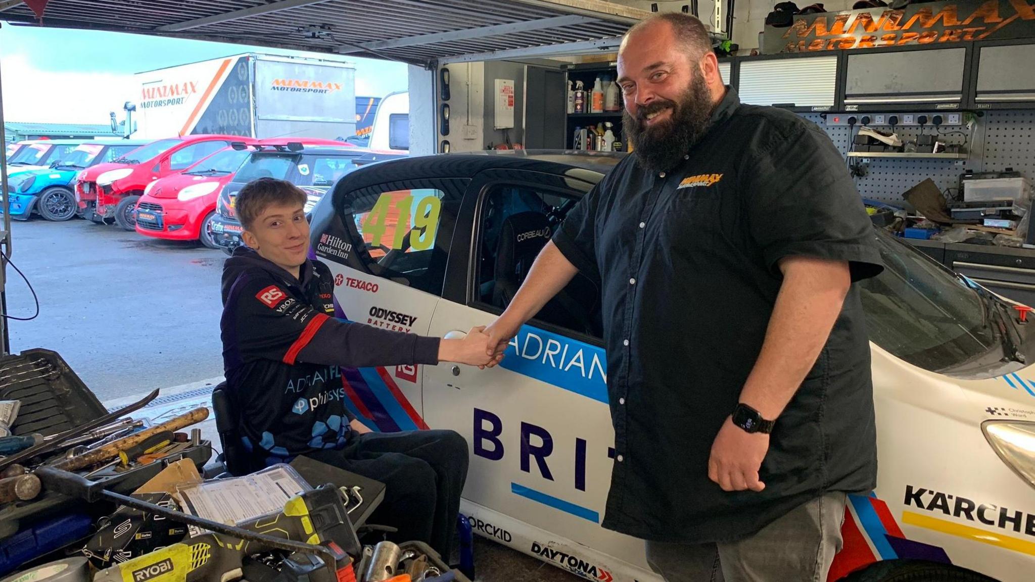 Ally Chalmers, a 17-year-old boy with short, blond hair, wearing a dark hoodie with sponsor names on it , shakes hands with engineer Scott MacKenzie who has a bald head and bushy beard. He is wearing a short-sleeved black shirt and jeans. They are smiling at the camera and standing in front of a racing car containing sponsor names