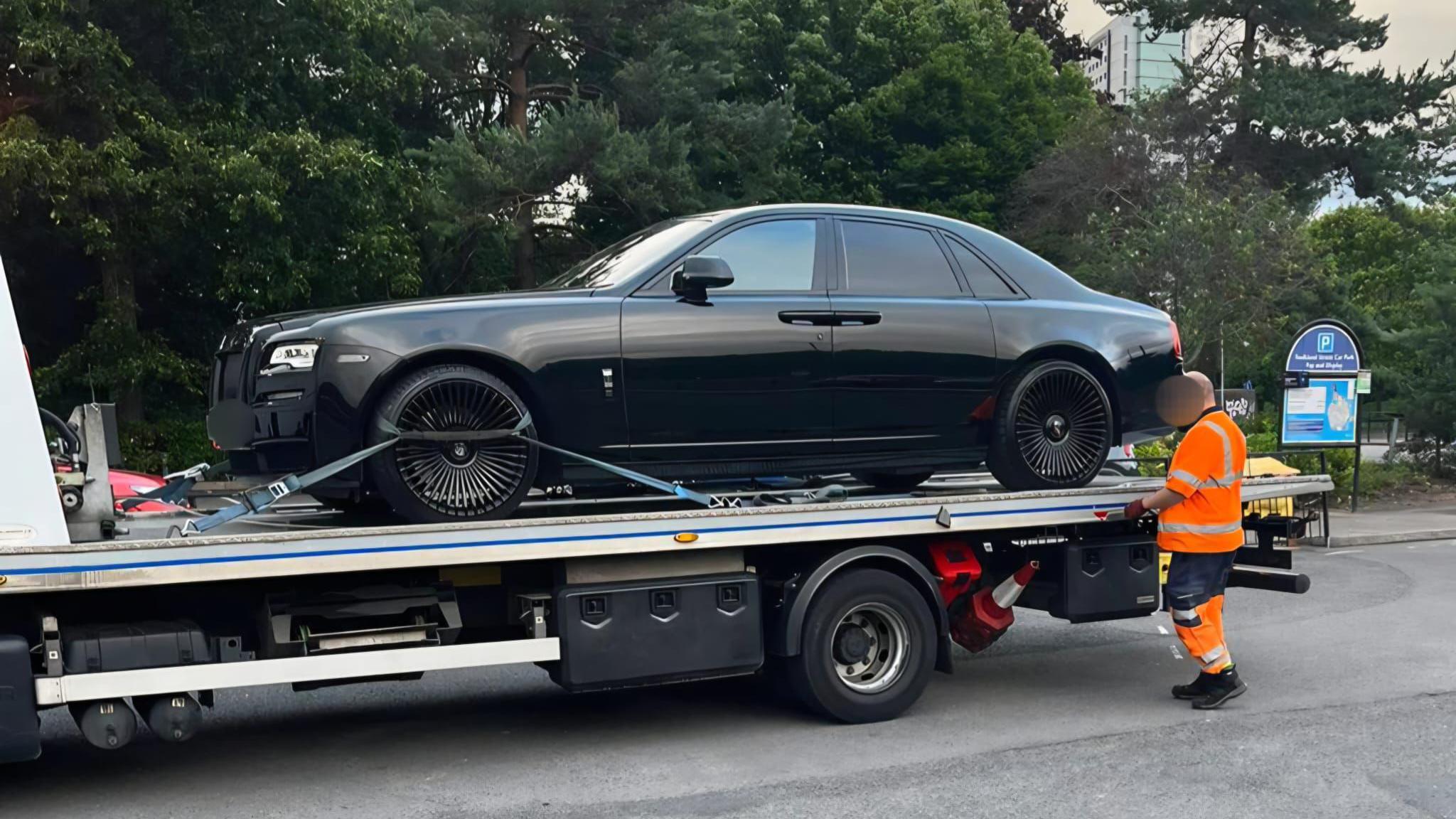 An expensive car on the back of a lorry. We can see a car park sign in the background. A man in a orange hi-vis uniform can be seen nearby, tightening the straps on the wheel of the car. We can see trees and a tower block in the background.