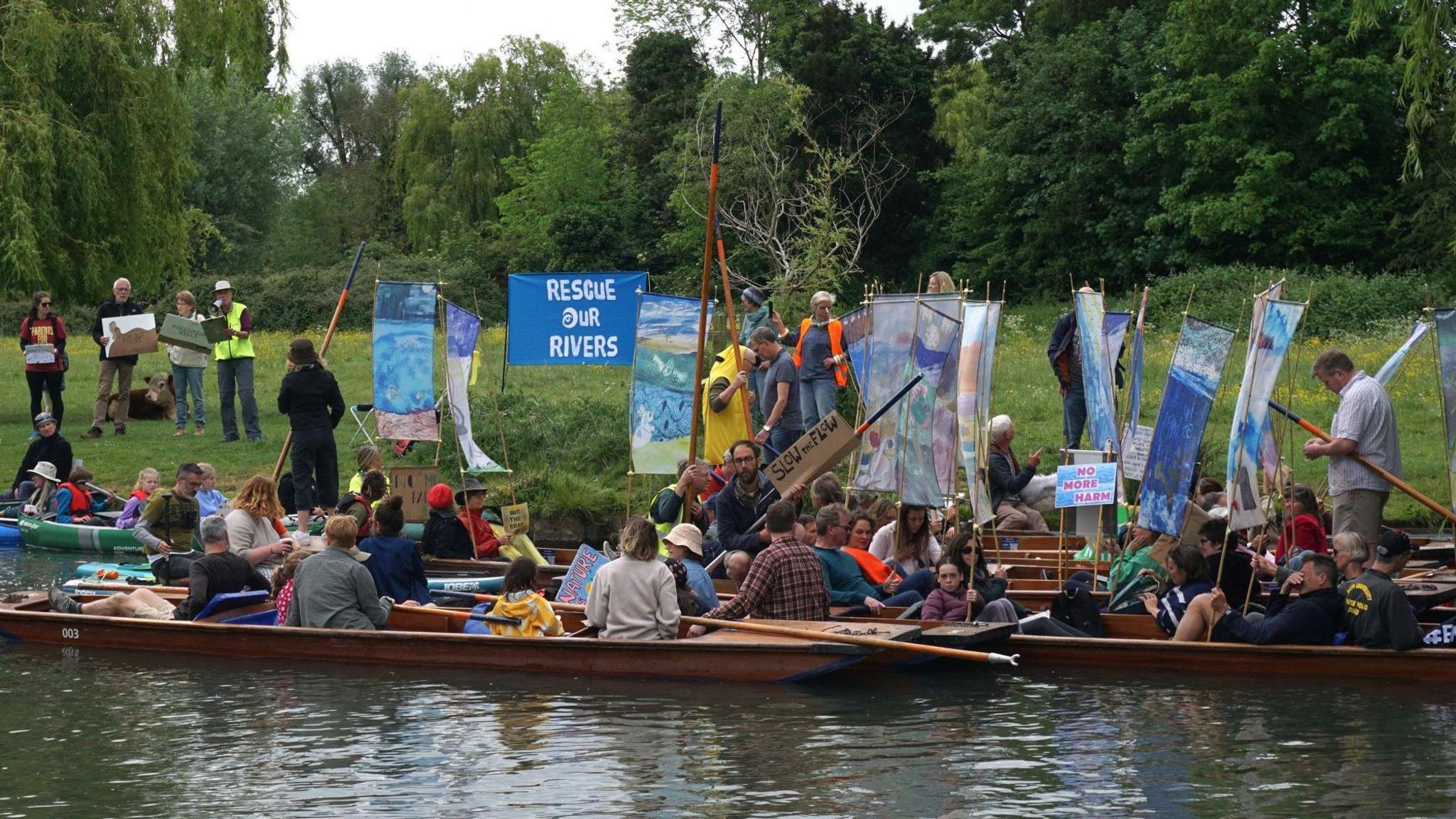 Several punts are clustered together at the river bank in Cambridge, next to a wildflower meadow lined by trees. They carry posters and flags with slogans such as "Rescue our Rivers" and "No more Harm". There are about 36 people in the photo including some children.