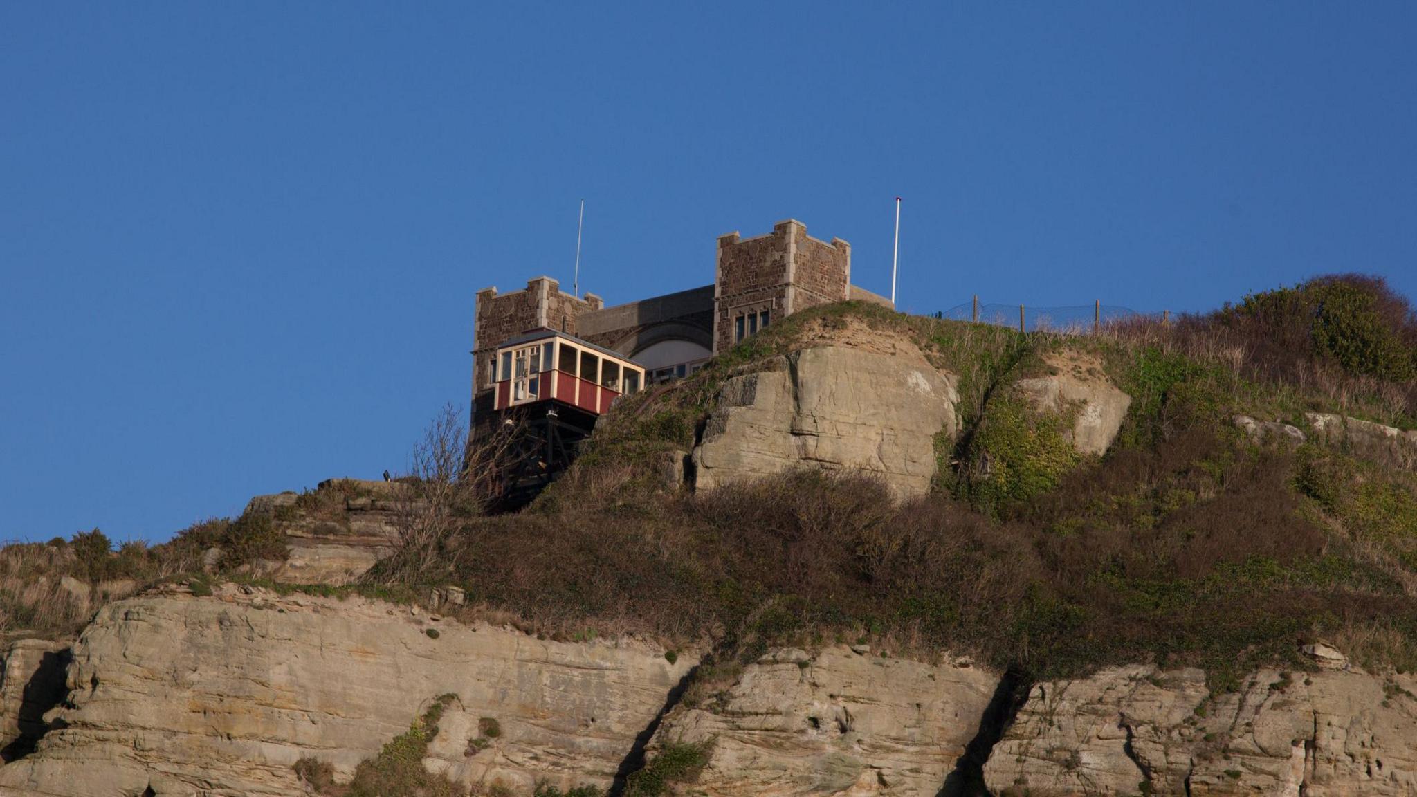 A fort-type building is sitting on top of a cliff. There is a yellow metal balcony on the front.
