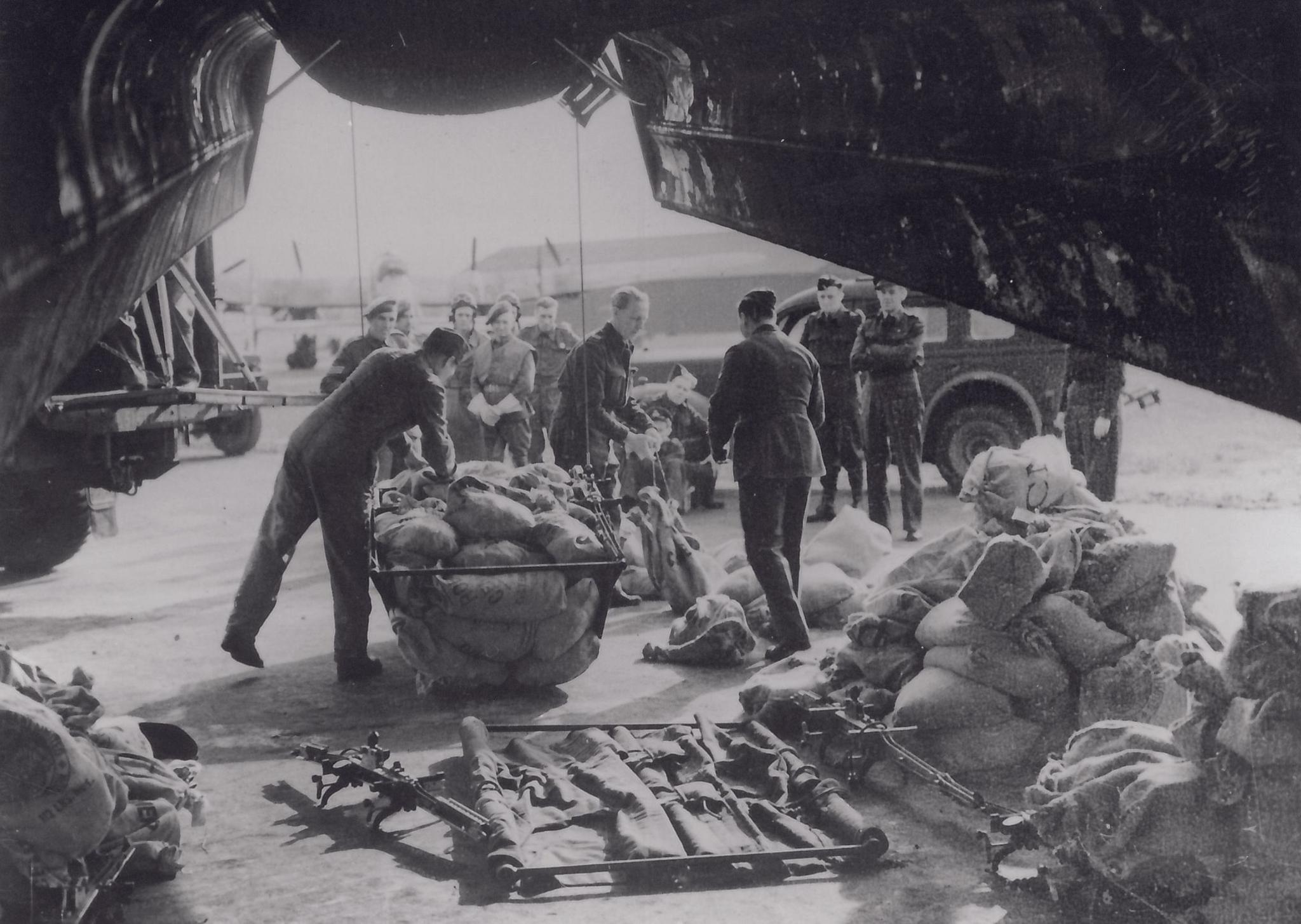A black and white photograph of a group soldiers loading sacks of food into a net that is being loaded on to a plane. The photo looks like it is taken from underneath or inside a plane. There are burlap sacks in heaps on the floor on the right of the photo. Vehicles can be seen in the background.