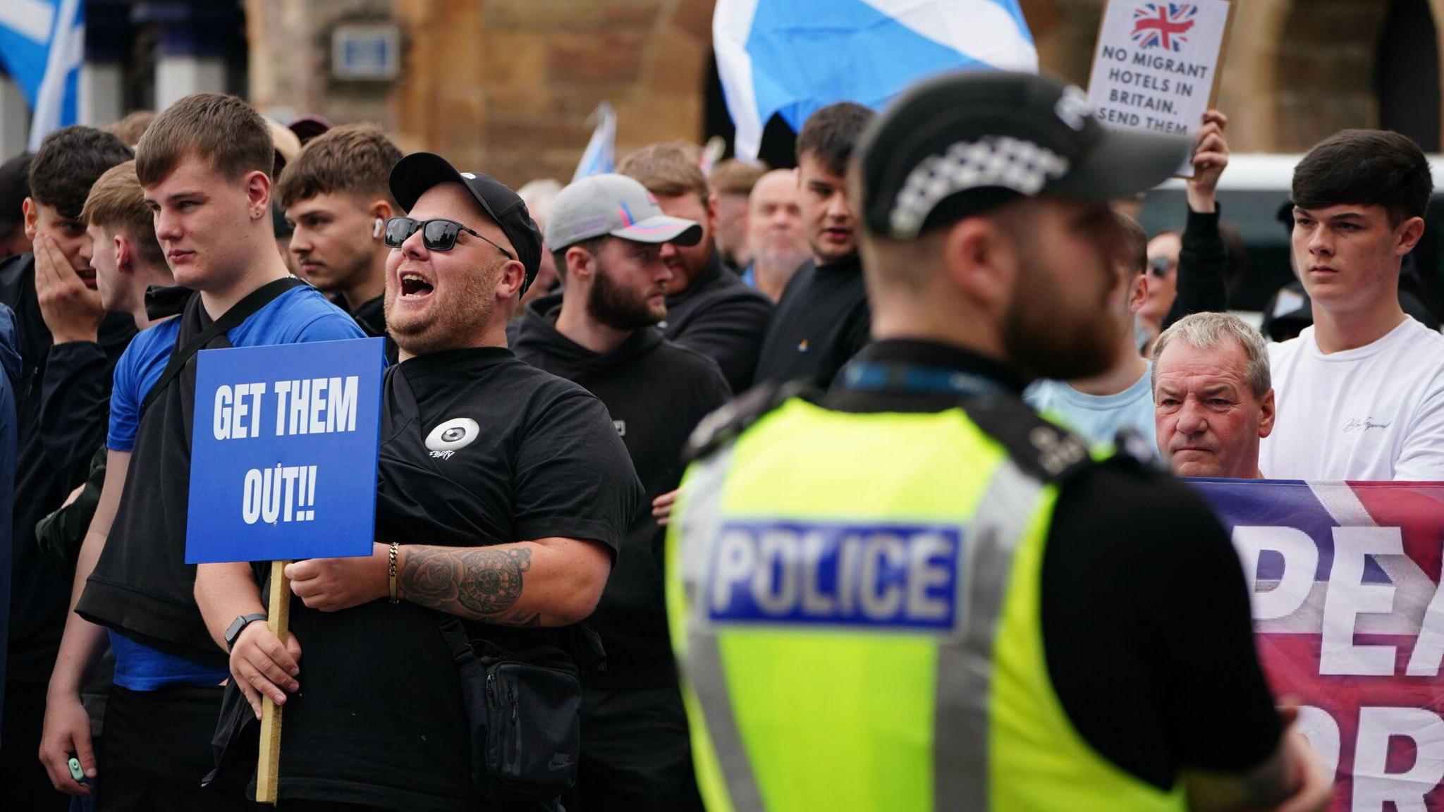 A group of men in dark clothing, one holds a blue sign which says "GET THEM OUT". A police officer faces the crowd and his high vis vest can be seen in the foreground