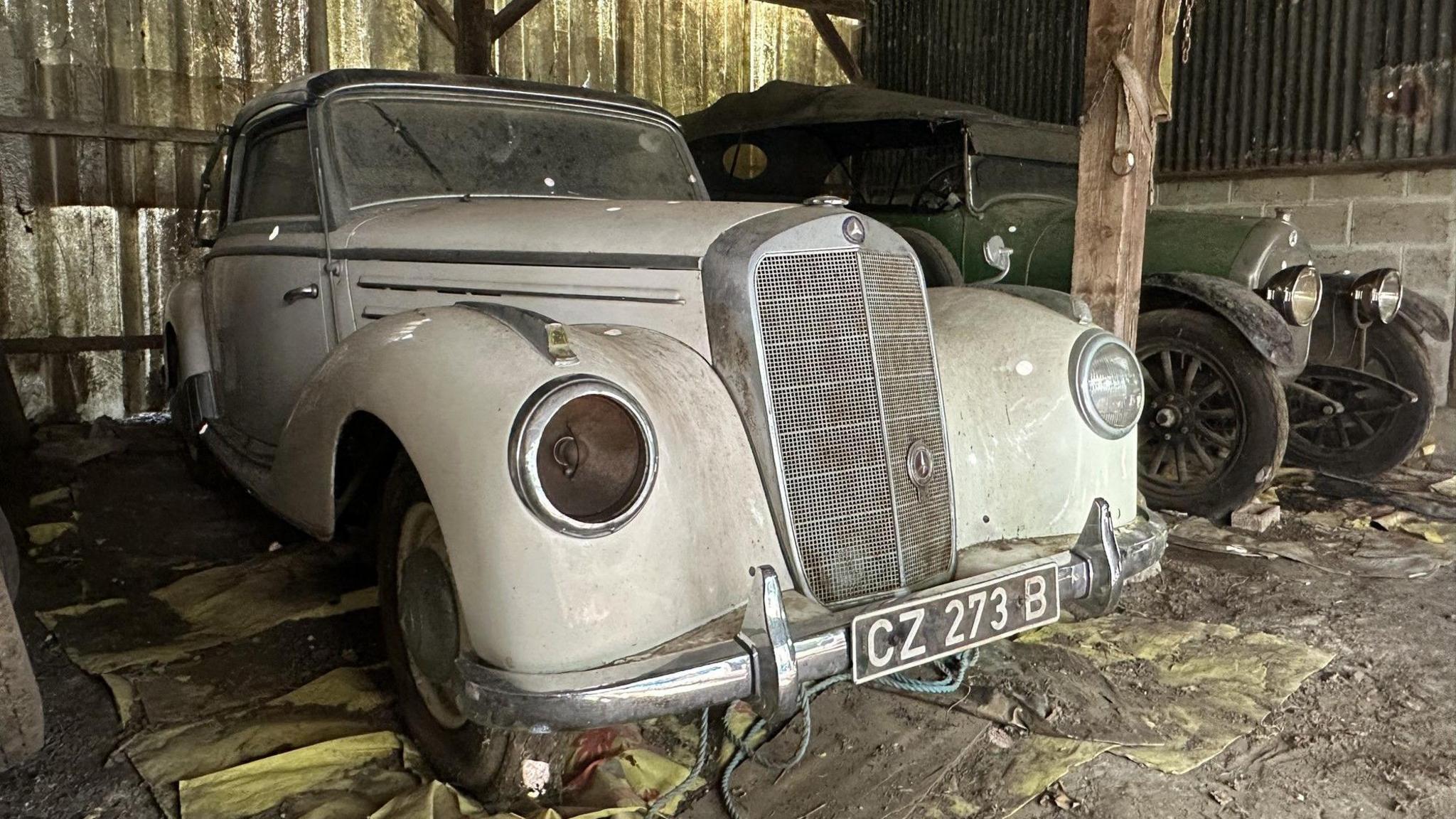 A dusty white Mercedes from the 1950s, with one headlight missing, next to a green 1921 Talbot open tourer in a barn 