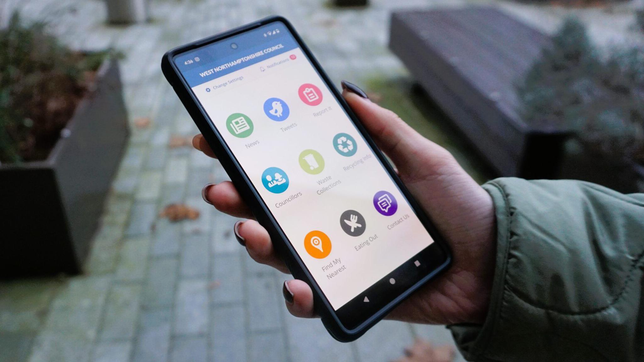 A hand holding a mobile phone. On the phone is the West Northamptonshire Council app with serval large icons.