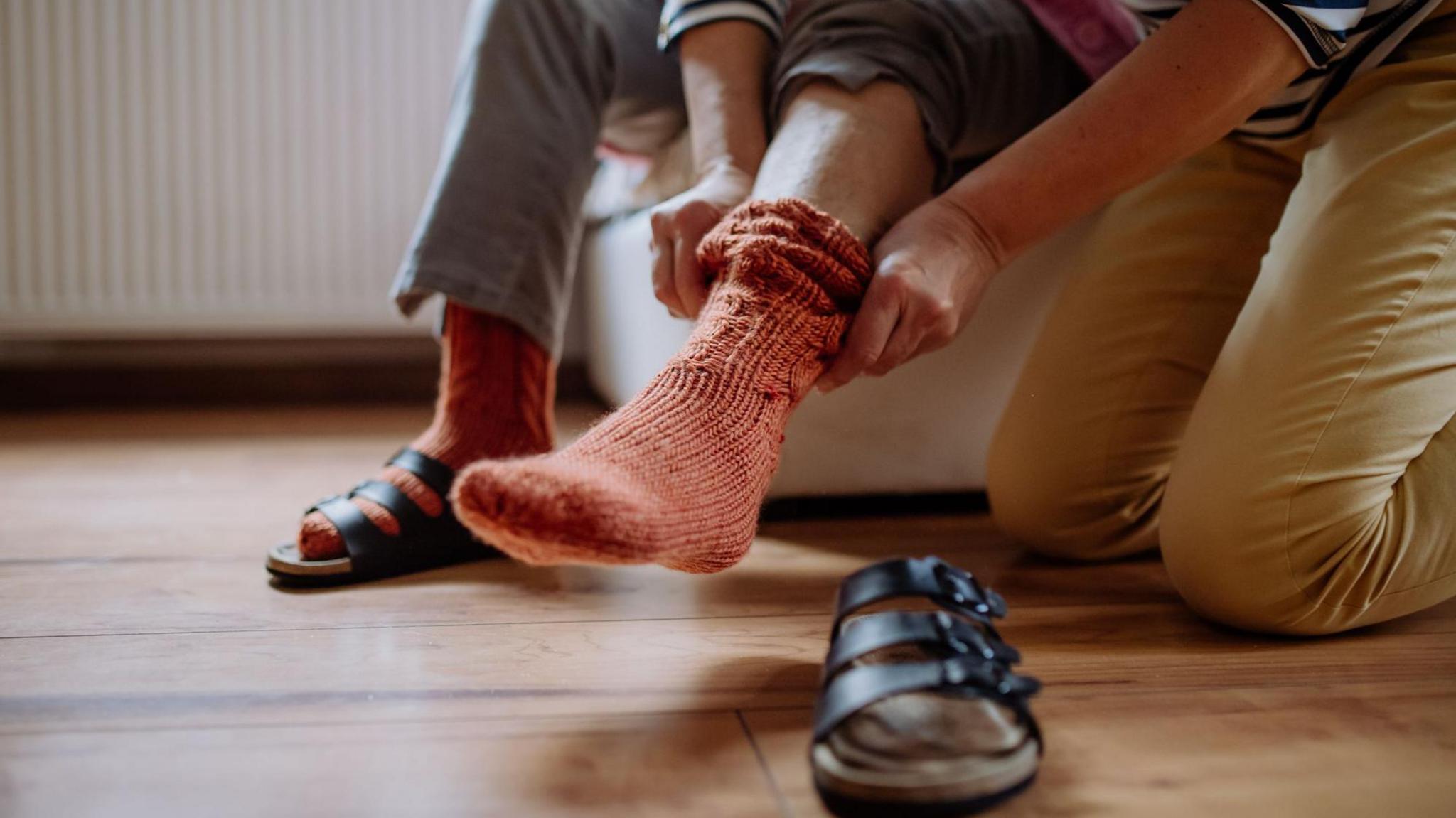 A stock image of a carer, who is kneeling on the floor, and an elderly person, who is sitting on a chair, put on their socks and sandals. The picture only pictures their arms, legs and the elderly person's feet.
