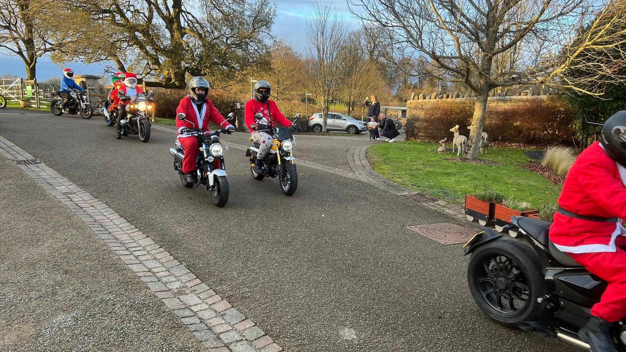 Five people in red Santa outfits seen driving down a quite road on a range of motorbikes, some of them small. There are a couple of people watching them pass, and two of them are riding side by side. 