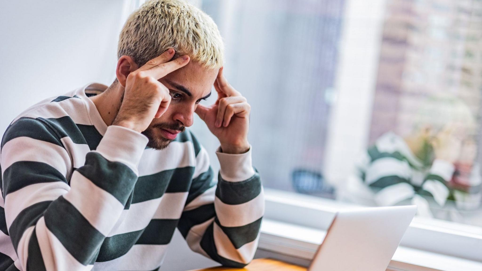 A young man with short bleached blonde hair and a beard, wearing a striped long-sleeved T-shirt, sits at a desk, looking toward an open laptop. He looks frustrated and is resting his fingers against the sides of the forehead.  A large window and houseplants are visible behind him. 