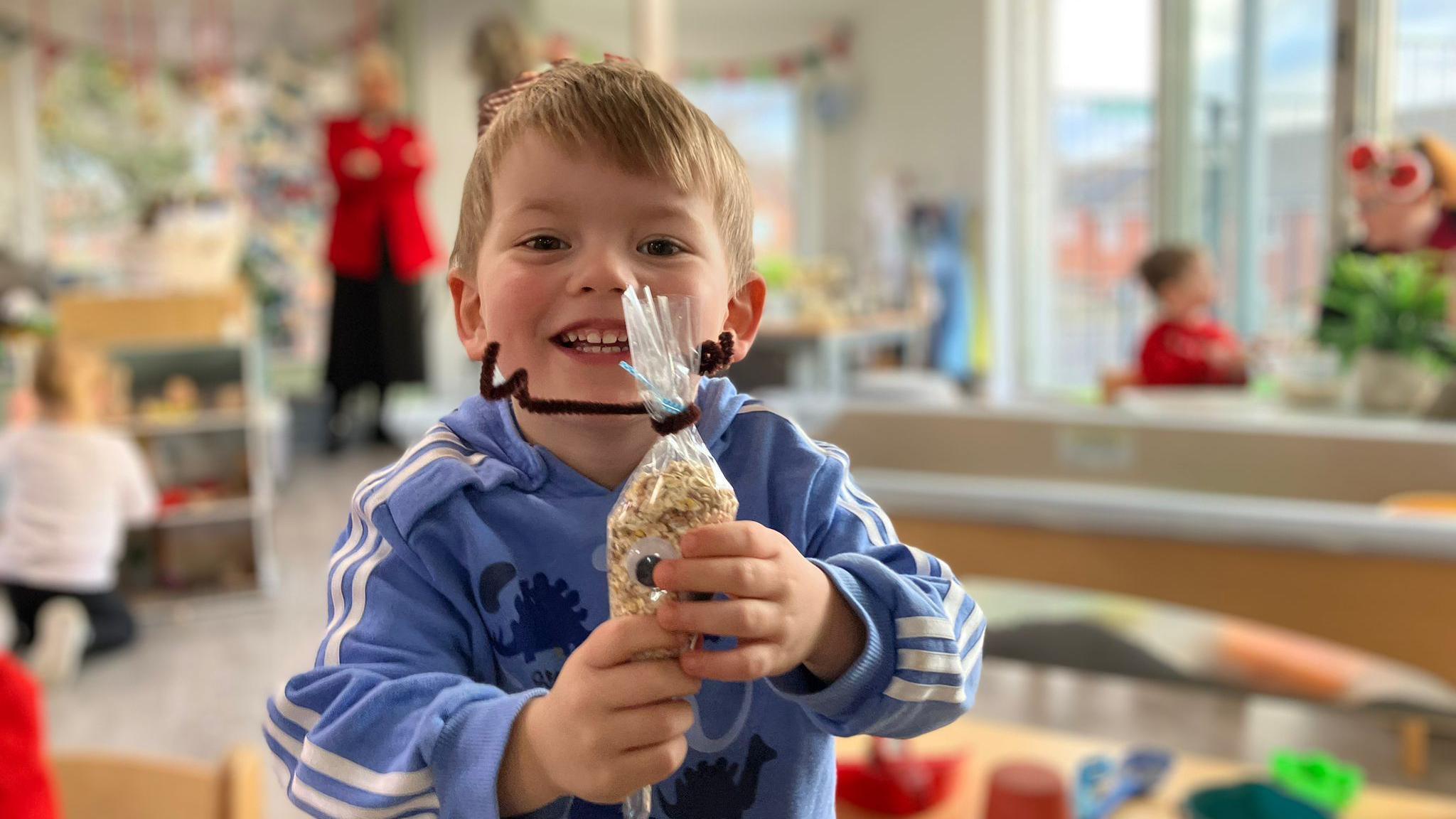 A child smiles at the camera as he holds up a craft he has made