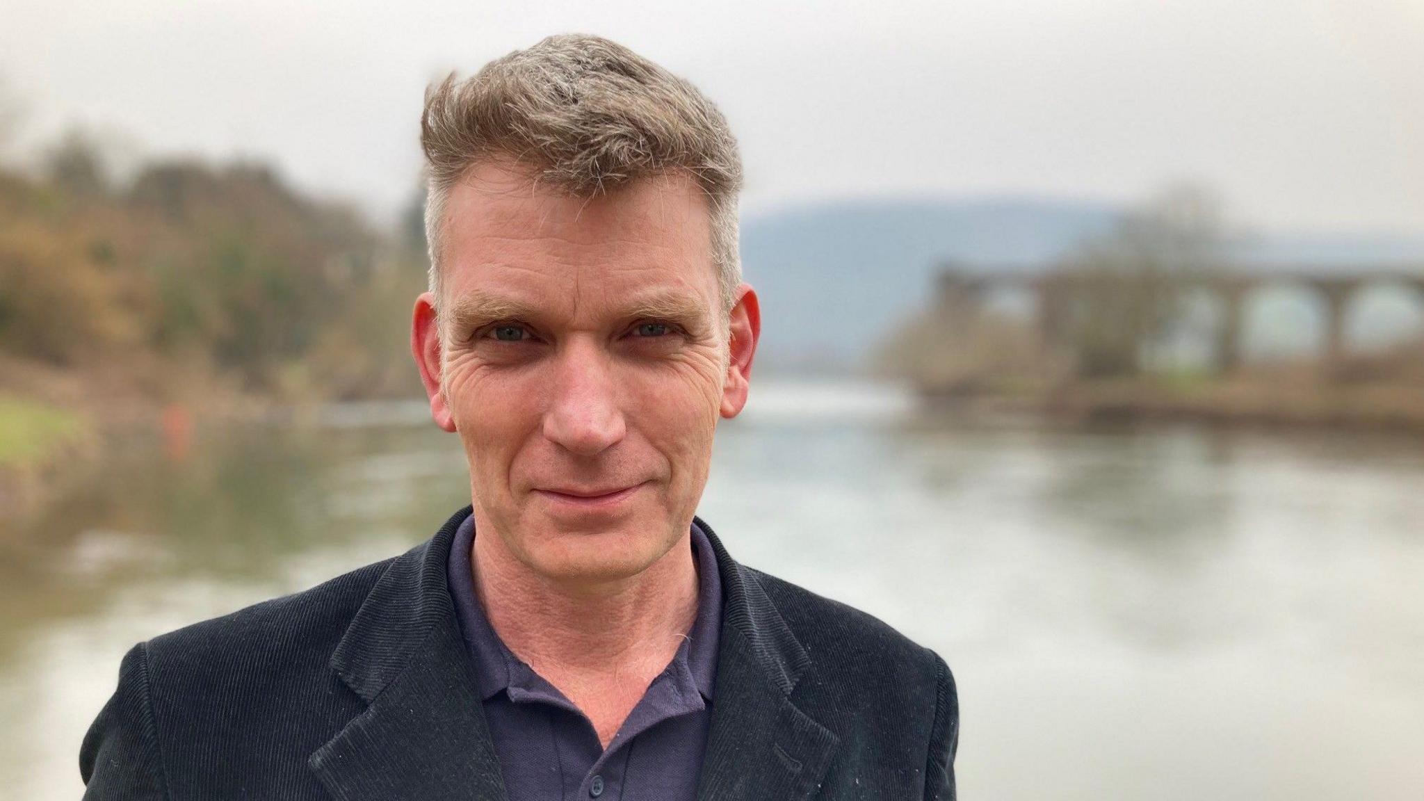 Simon Evans, chief executive officer of the Wye and Usk Foundation stands looking at the camera in front of the river Wye at Monmouth in a black jacket and purple shirt.